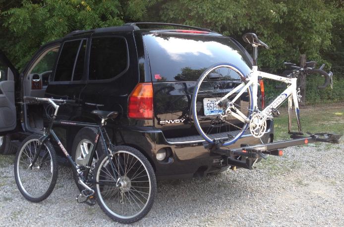 A black SUV parked on gravel with a bike rack attached to the back. One bicycle is secured to the rack, while another bicycle is leaning against the side of the vehicle. The vehicle's rear doors are open, indicating that it is in use. Lush greenery is visible in the background.