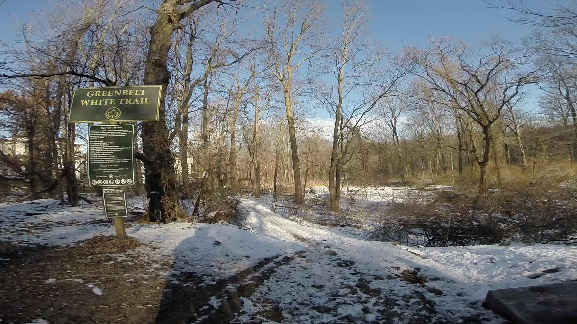 Image of a snowy trail in a forest setting, featuring a sign for the Greenbelt White Trail. The sign provides information about the trail and its features, while the surrounding landscape includes bare trees and snow-covered ground, with a clear blue sky in the background. Richmond Avenue and Forest Hill road mountain bike trail.