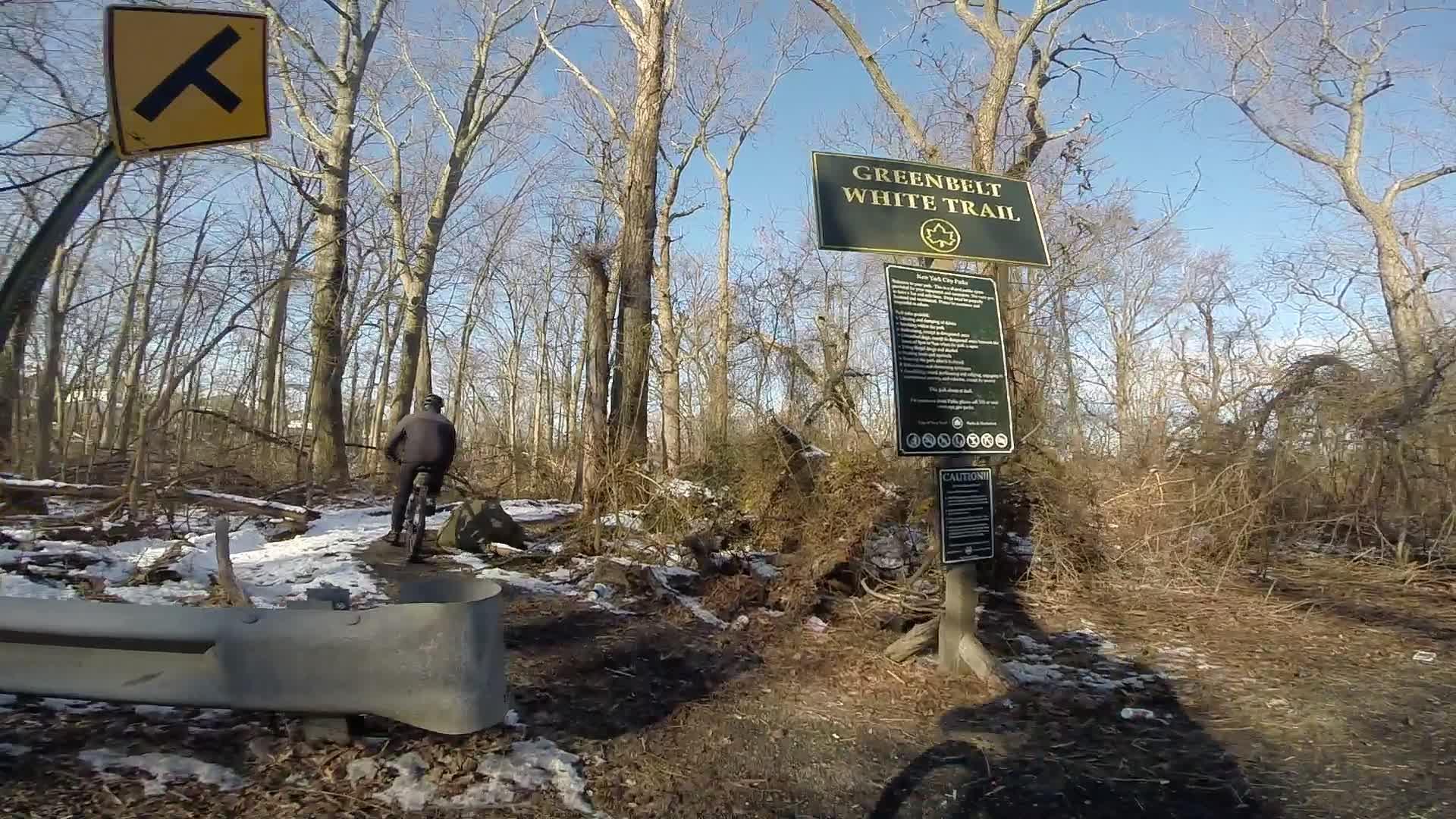 A cyclist rides along a snowy path in a wooded area, with a sign for the Greenbelt White Trail visible nearby. The scene shows bare trees and a clear blue sky, creating a peaceful outdoor atmosphere. Richmond Avenue and Forest Hill road mountain bike trail.