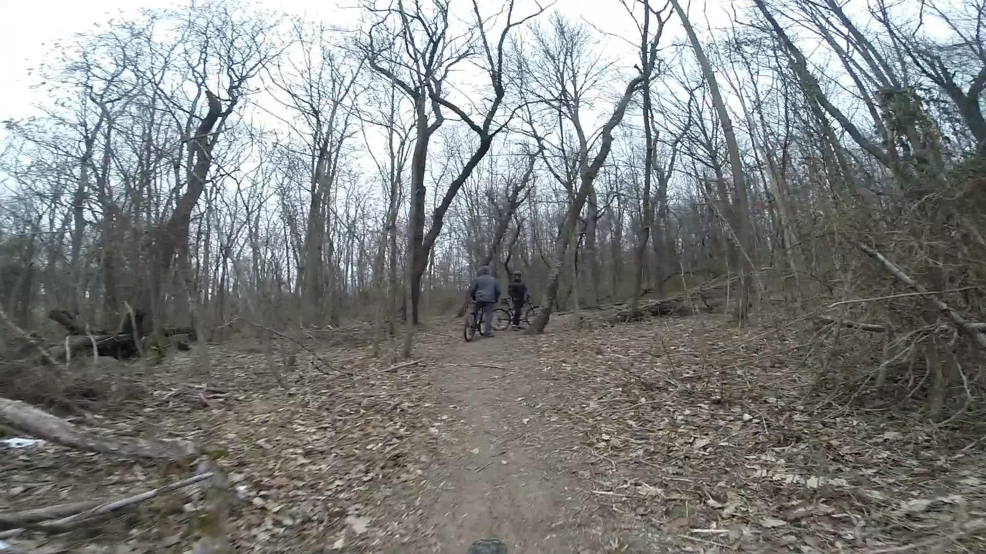 Two mountain bikers pause on a dirt trail in a wooded area during early spring, surrounded by leafless trees and scattered leaves on the ground. The atmosphere is calm and quiet, suggesting a cool, overcast day. Richmond Avenue and Forest Hill road mountain bike trail.