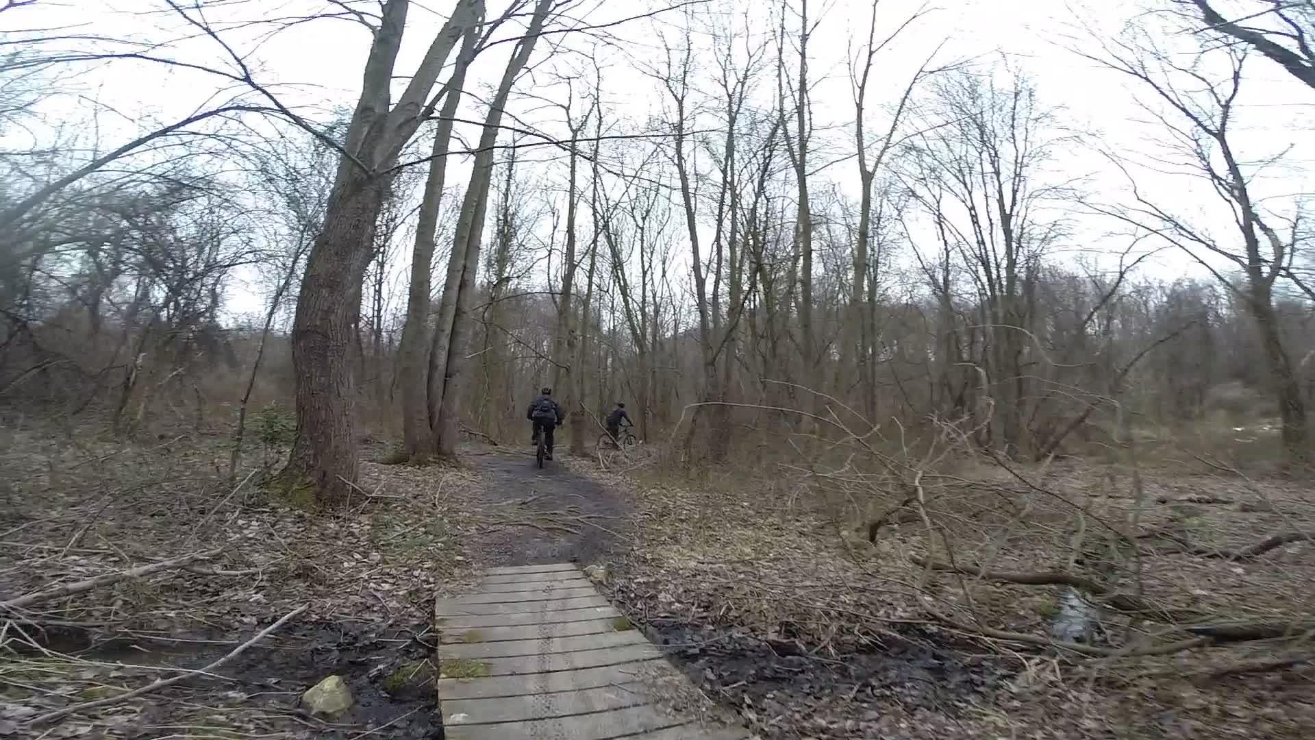 Two mountain bikers riding along a narrow, muddy trail through a wooded area with bare trees. A wooden bridge crosses a small stream, surrounded by dried leaves and branches. The scene is overcast, suggesting an early spring or late fall atmosphere. White Clay Creek mountain bike trail.