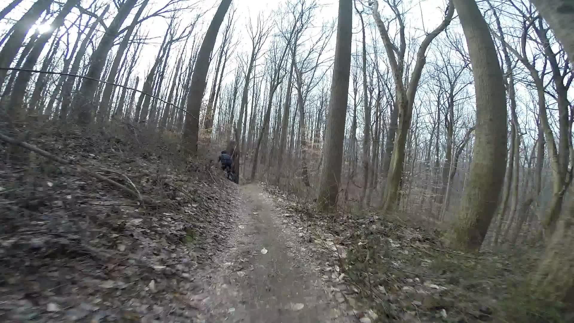 A person riding a mountain bike along a narrow dirt trail in a wooded area during late autumn or winter, surrounded by bare trees and scattered leaves on the ground. White Clay Creek mountain bike trail.