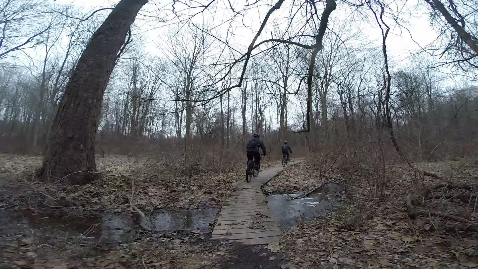 Two mountain bikers riding along a narrow wooden path through a dense, leafless winter forest. The path crosses over a small stream, surrounded by bare trees and fallen leaves, under a cloudy sky. White Clay Creek mountain bike trail.