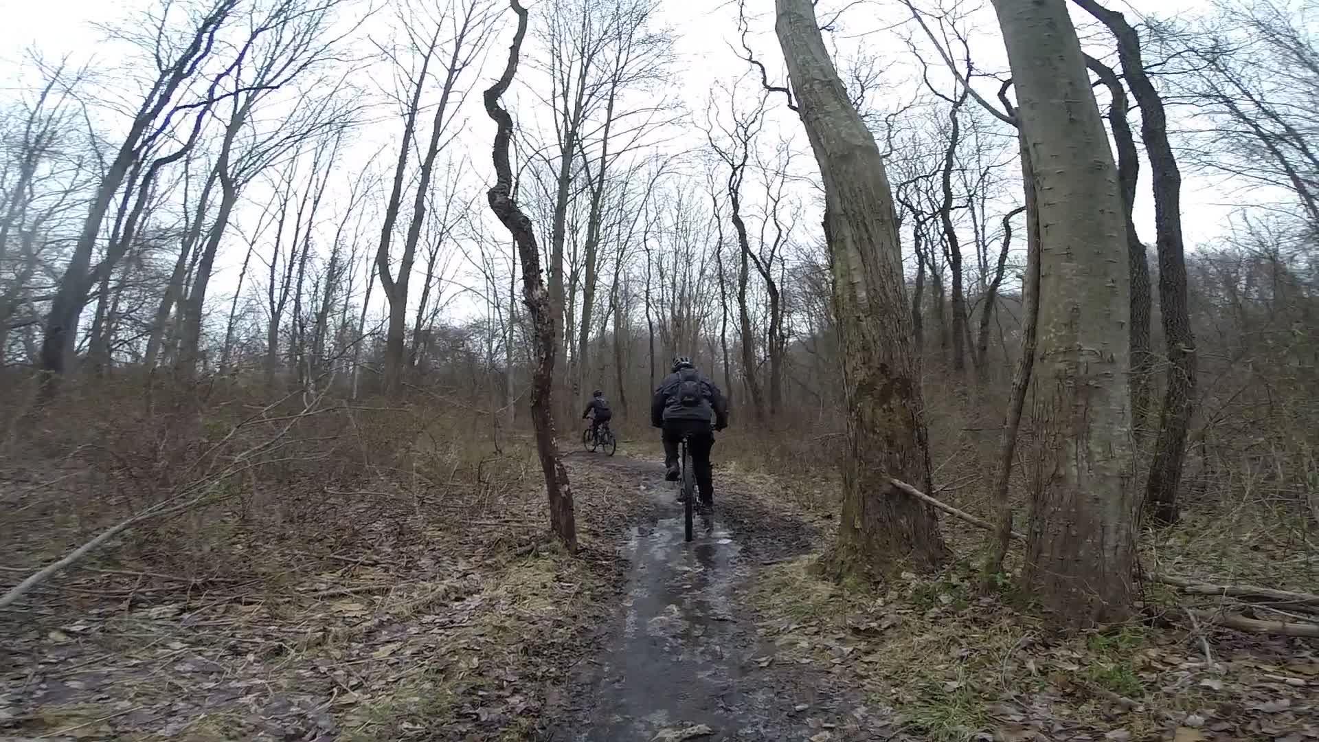 Two mountain bikers riding on a muddy, narrow trail through a wooded area during an overcast day. The scene features leafless trees and sparse vegetation, with the riders appearing in the distance, surrounded by the natural landscape. White Clay Creek mountain bike trail.