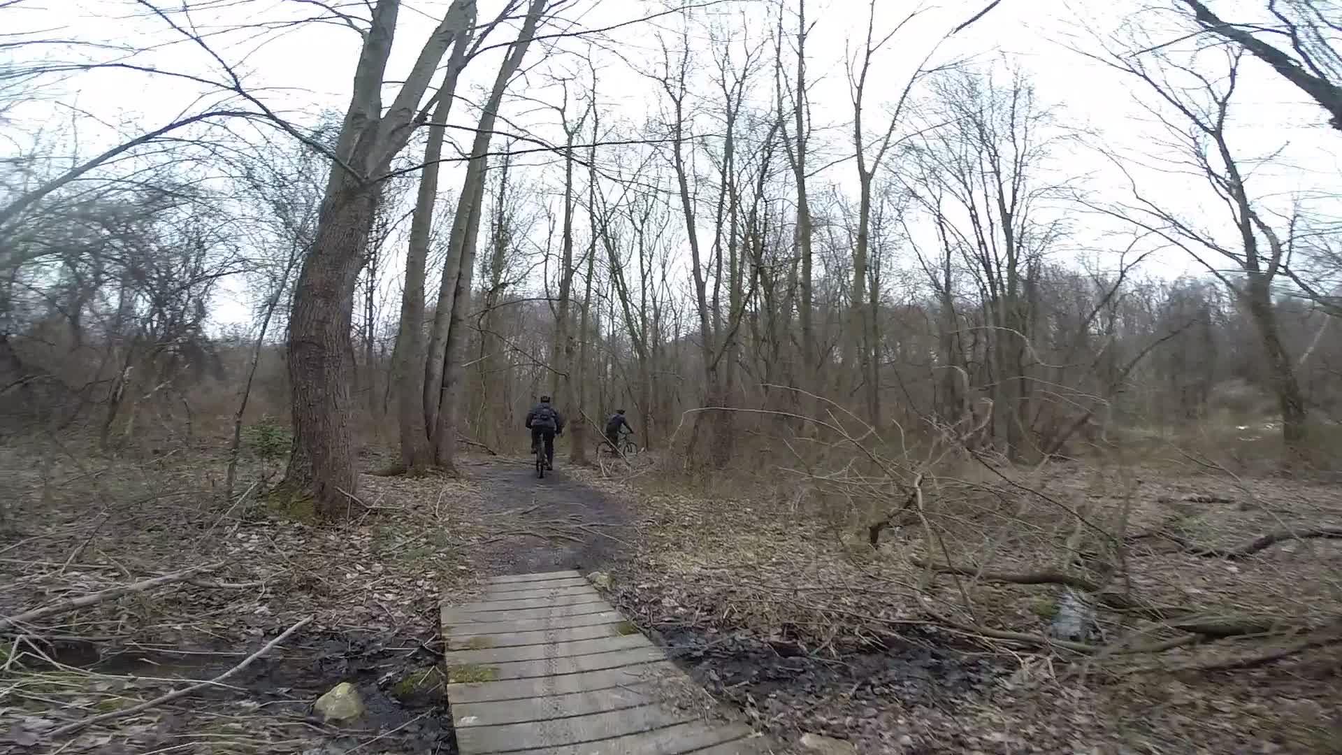Two cyclists riding on a dirt trail through a wooded area with bare trees and sparse vegetation, crossing a wooden bridge over a small stream. The scene is set in early spring or late winter, with a cloudy sky overhead. White Clay Creek mountain bike trail.