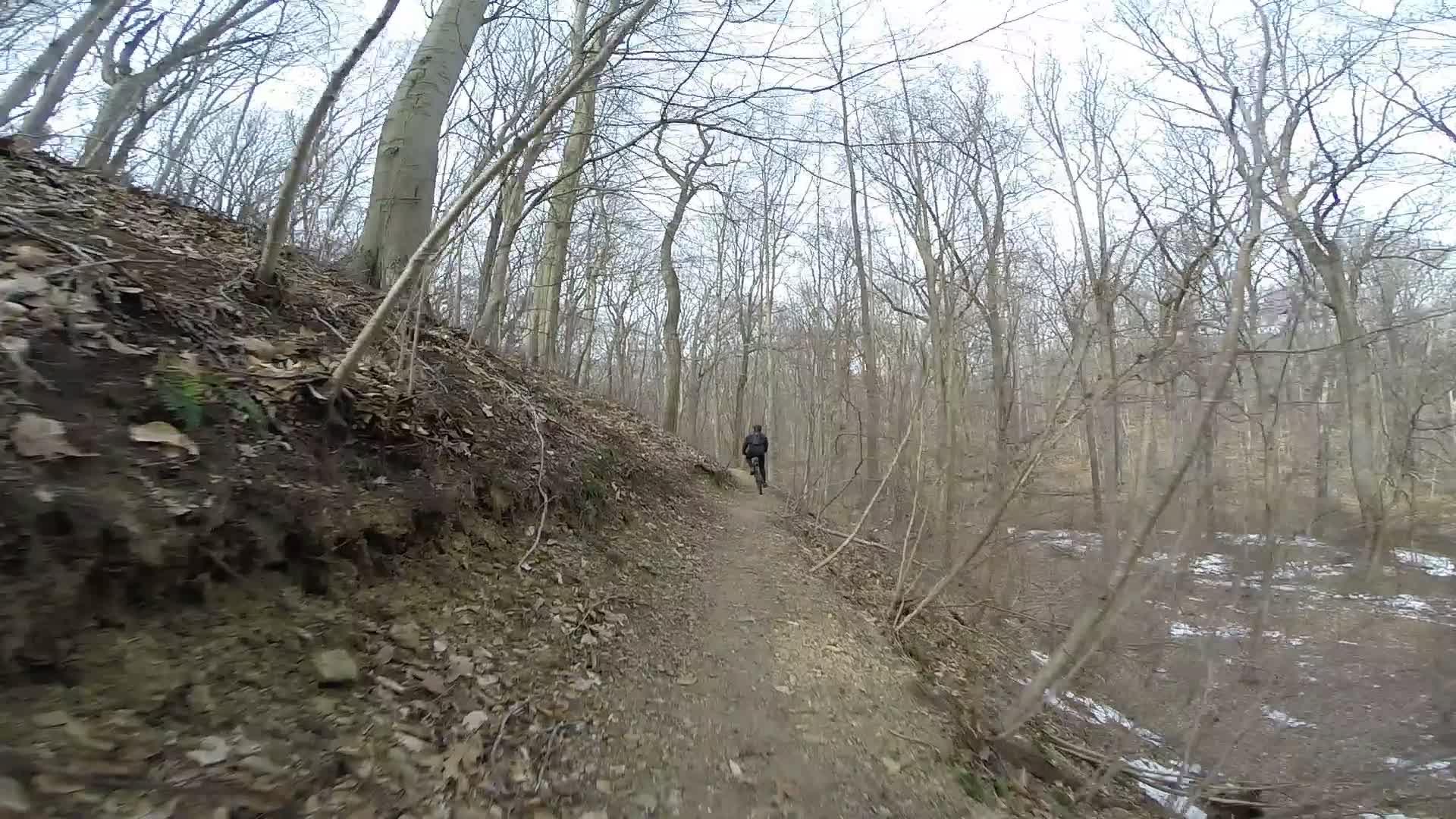 A person walking along a dirt path in a wintery forest, surrounded by bare trees and scattered leaves. The trail is bordered by a slight incline on one side and a view of a creek or river on the other, with patches of snow visible on the ground. The scene is overcast, indicating a cool, cloudy day. White Clay Creek mountain bike trail.