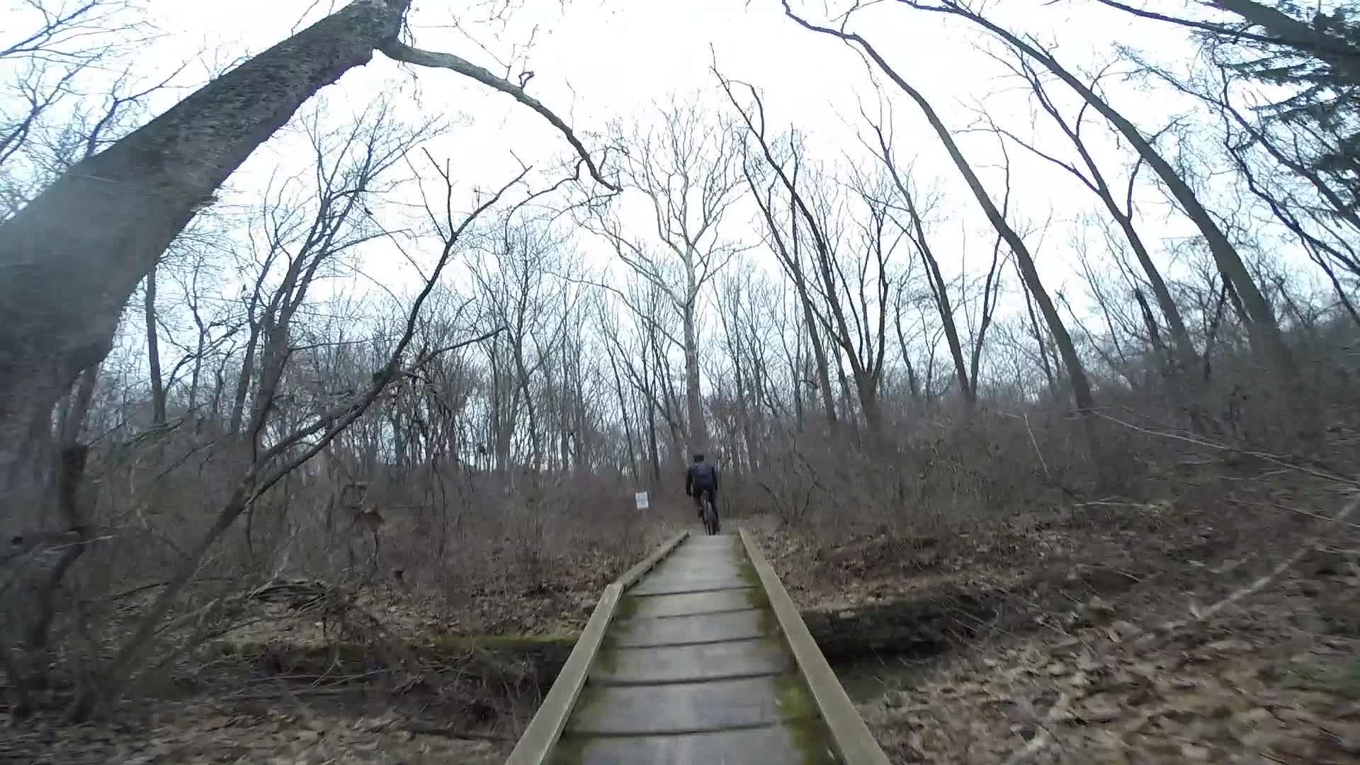 A mountain biker rides along a wooden bridge through a wooded area, with bare trees and brown foliage surrounding the path. The scene is set on a cloudy day, creating a serene and nature-focused atmosphere. White Clay Creek mountain bike trail.