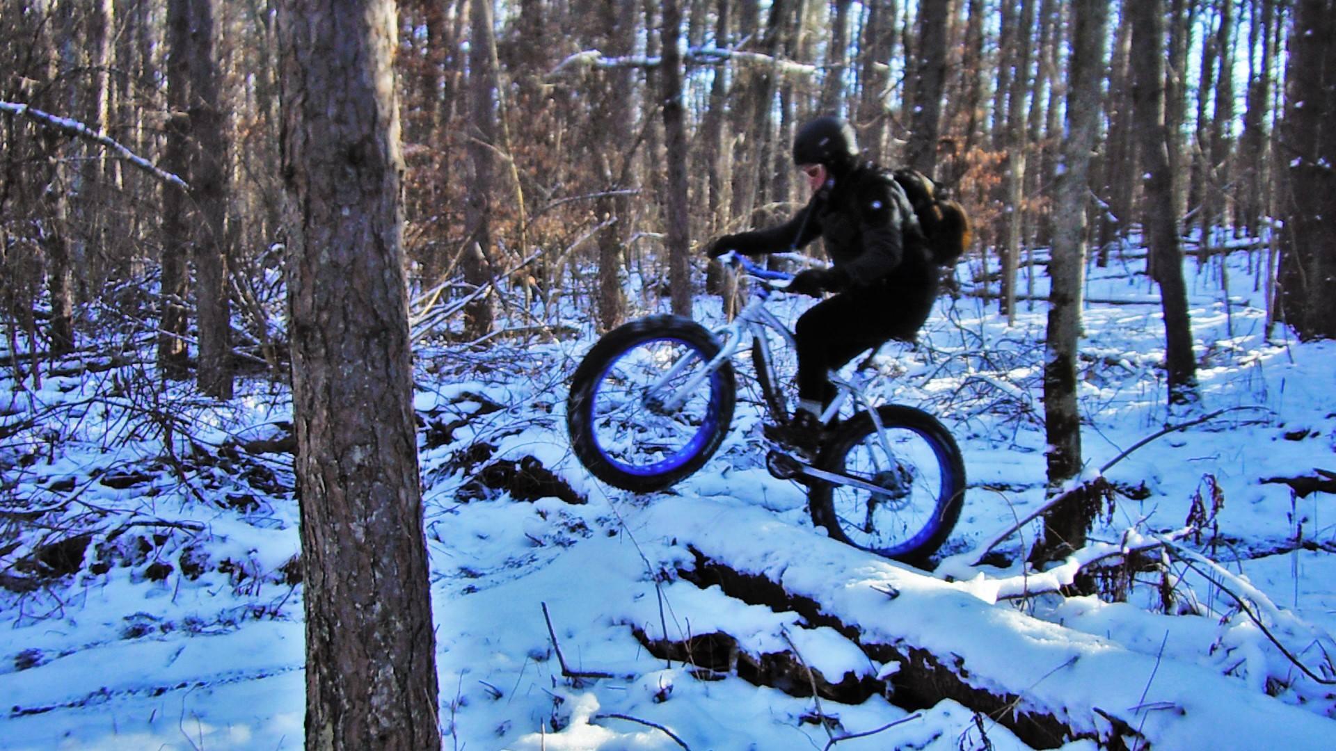 A person riding a fat bike over a snowy trail in a forest. The cyclist is wearing a helmet and winter gear, with trees and snow-covered ground surrounding them. Colgate mountain bike trail.