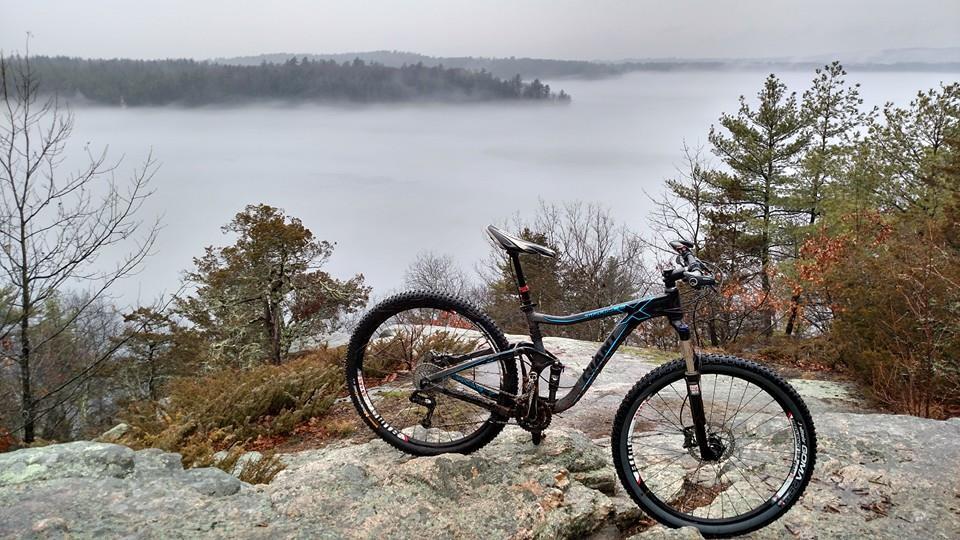 A mountain bike resting on a rocky outcrop, overlooking a foggy landscape with trees in the background. The scene conveys a tranquil outdoor setting, featuring muted colors and a sense of exploration. Depot Road Singletracks/FOMBA mountain bike trail.