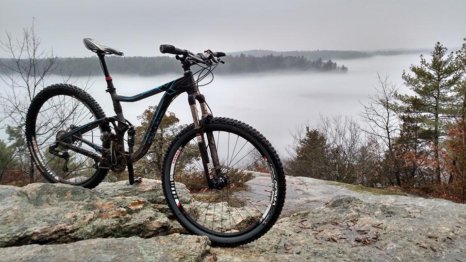 A mountain bike leaning against a rock with a foggy lake and forested landscape in the background. The scene captures a serene, overcast day, highlighting the natural beauty of the outdoors. Depot Road Singletracks/FOMBA mountain bike trail.