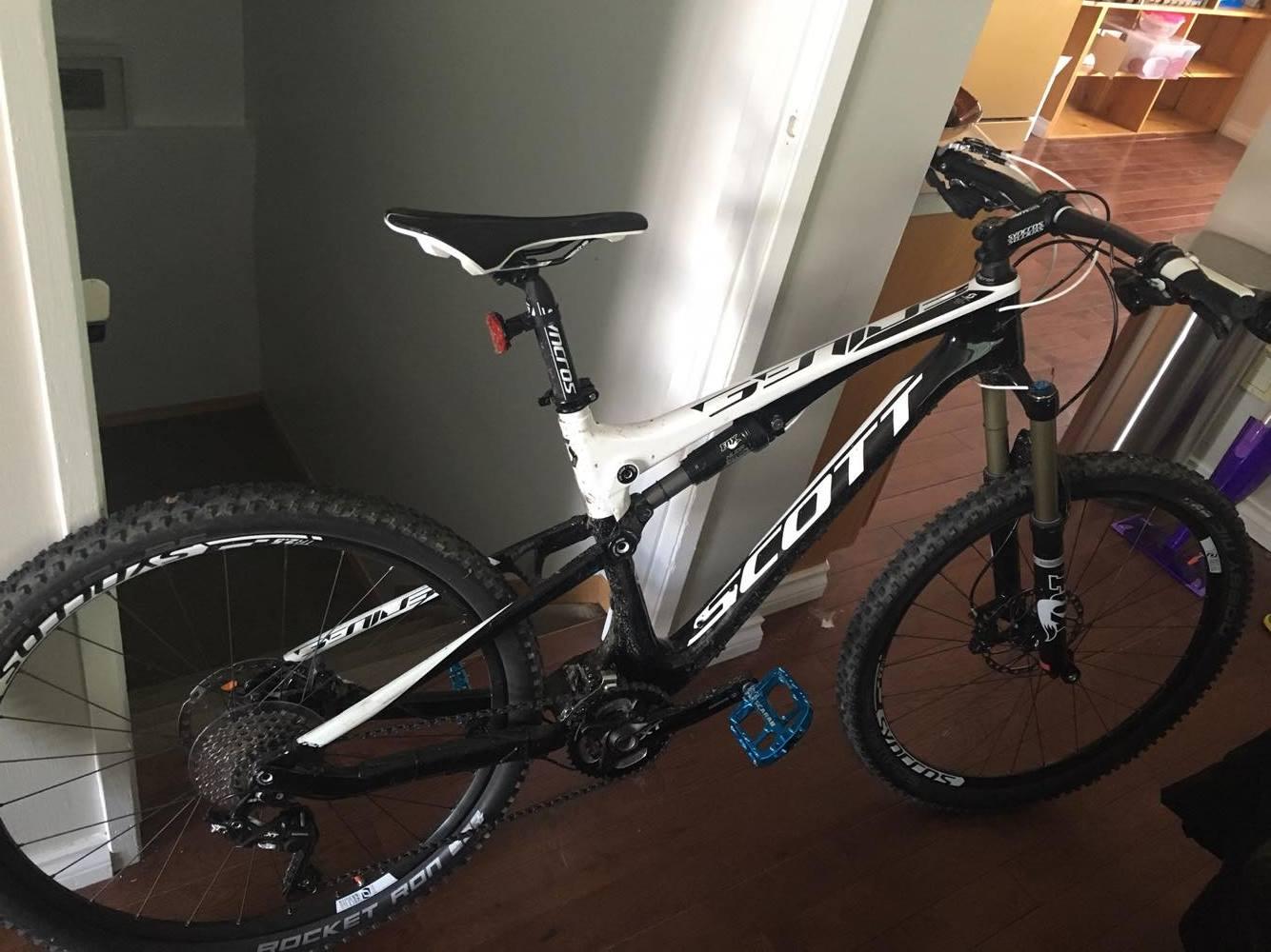 Scott Genius 720: A black and white mountain bike is positioned inside a hallway, leaning against the wall. It features sturdy tires designed for off-road biking, with a prominent "Scott" logo on the frame. The bike's seat is adjustable, and it has a visible gear system at the back. In the background, a glimpse of a living space with wooden flooring and some furniture can be seen.