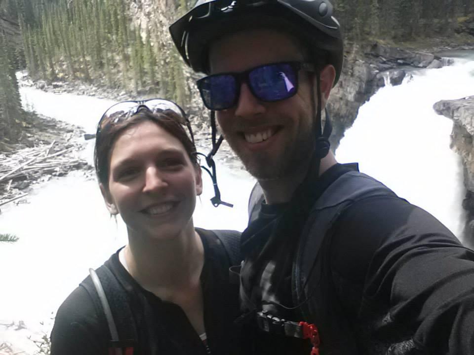 A smiling couple taking a selfie outdoors near a rushing river, surrounded by tall green trees and rocky terrain. The man is wearing sunglasses and a helmet, while the woman has sunglasses and a carefree expression. Snake Indian Falls mountain bike trail.