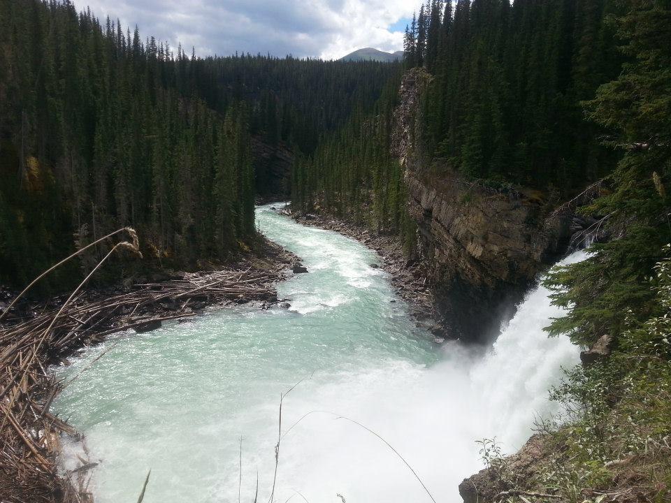 A scenic view of a river flowing through a dense forest, with vibrant green trees lining both sides. The river features a turquoise hue and cascades over a rocky ledge, creating a waterfall. In the background, mountains are visible under a partly cloudy sky. Debris and fallen branches are scattered along the riverbank, enhancing the natural landscape. Snake Indian Falls mountain bike trail.