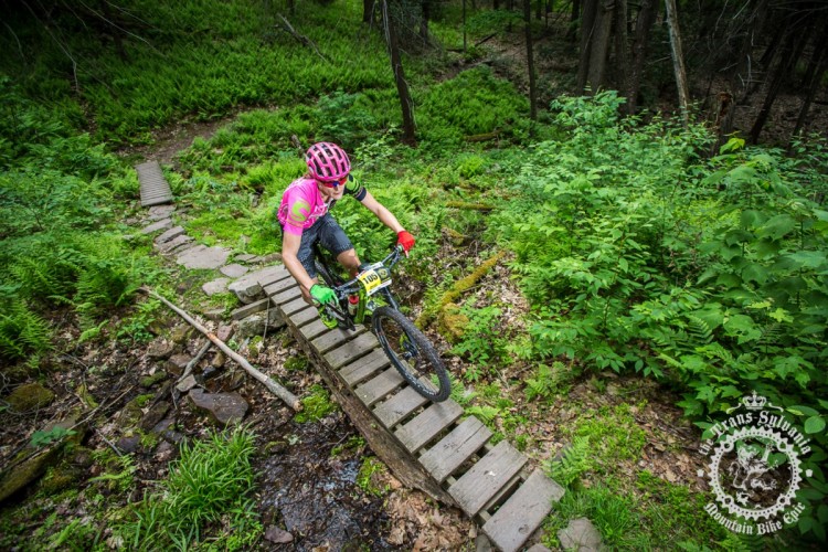 A mountain biker in a bright pink shirt navigates a wooden bridge while riding through a lush, green forest. The background features dense foliage and a stream, creating a vibrant outdoor scene.