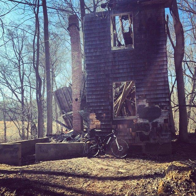 A dilapidated, partially burned house stands among leafless trees, with visible damage to its structure. A black bicycle is leaning against a concrete foundation in the foreground, while remnants of the building, including a brick chimney and broken windows, can be seen. The scene is illuminated by sunlight, highlighting the surrounding forest. Patapsco Valley State Park (Avalon Area) mountain bike trail.