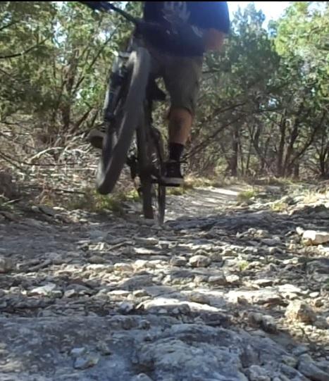 A mountain biker performing a jump over a rocky trail surrounded by trees. The bicycle's front wheel is lifted off the ground while the rider’s legs are extended. The scene captures the action and rugged terrain typical of off-road biking. Madrone Trail mountain bike trail.