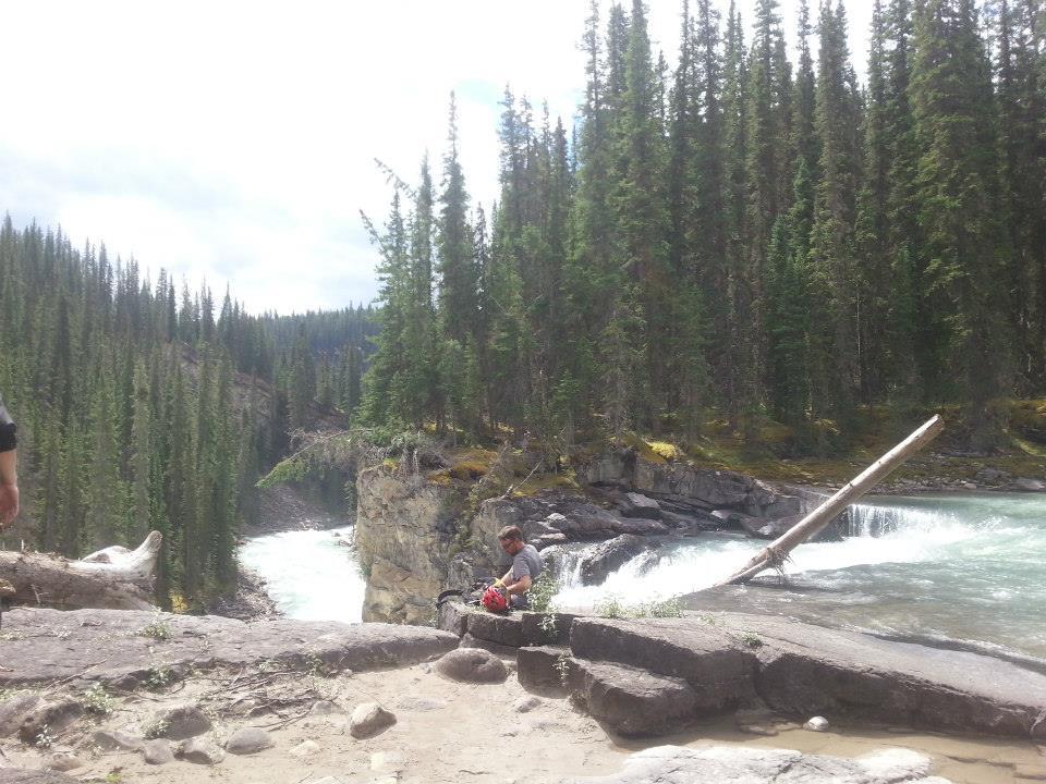 A scenic view of a river flowing through a forested area, with tall pine trees lining the banks. In the foreground, a person is seated on a rocky outcrop near the water, while another individual stands nearby. The sky is partly cloudy, and the water cascades gently over rocks, creating a tranquil atmosphere. Snake Indian Falls mountain bike trail.