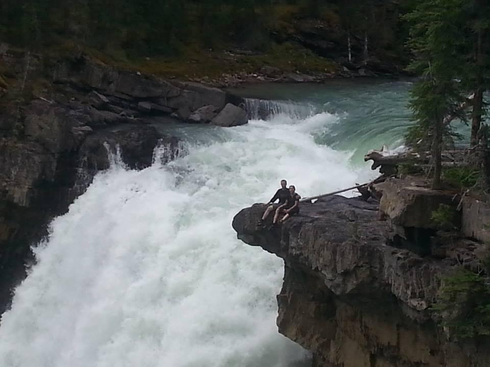 Two people sitting on the edge of a rocky outcrop overlooking a powerful waterfall, surrounded by lush greenery. The water cascades dramatically into a turquoise pool below, creating a misty ambiance. Snake Indian Falls mountain bike trail.