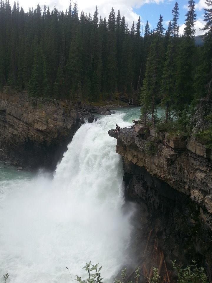 A breathtaking view of a waterfall cascading over a rocky ledge, surrounded by dense evergreen trees. The water is rushing down with significant force, creating mist and foam at the base. The scene captures the natural beauty of a wilderness landscape under a partly cloudy sky. Snake Indian Falls mountain bike trail.