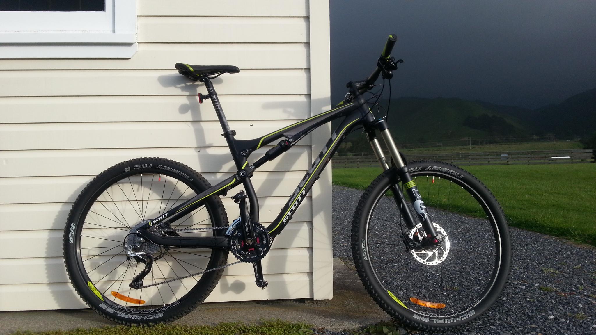 Scott Genius 720: A mountain bike with a black frame accented by green highlights, leaning against a beige wooden wall. The bike features front suspension, wide tires with tread patterns, and disc brakes. In the background, there is a grassy area and dark storm clouds approaching the mountains.