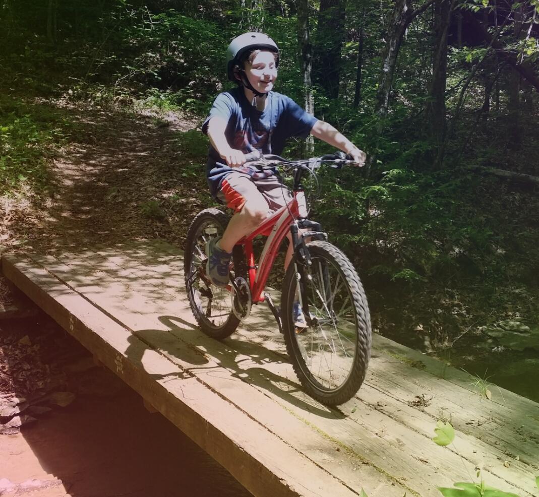 A young boy wearing a helmet rides a red bicycle across a wooden bridge in a wooded area, smiling as he enjoys the outdoors. Sunlight filters through the trees, casting dappled light on the trail. Sheltowee Trace - Laurel Lake Trail mountain bike trail.