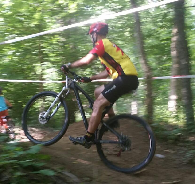 A mountain biker in a red and yellow jersey performs a wheelie on a dirt trail surrounded by greenery. The rider is focused and wearing a helmet, with a second cyclist visible in the background. The image conveys motion and excitement in a natural outdoor setting. England Idlewild Mountain Biking Park mountain bike trail.