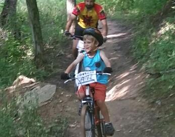 A young boy in a blue tank top and orange shorts joyfully rides a bicycle on a dirt trail surrounded by trees, while an adult cyclist in a colorful jersey follows closely behind. England Idlewild Mountain Biking Park mountain bike trail.