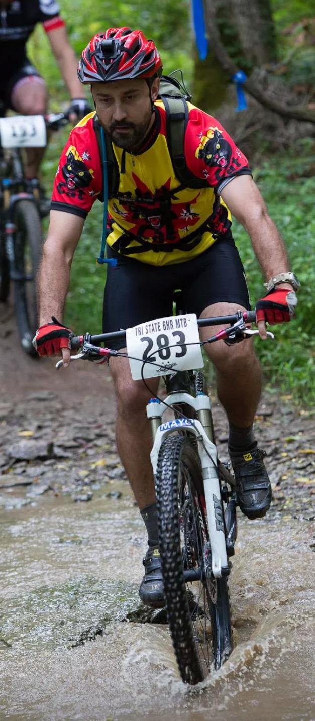 A mountain biker navigates through a muddy trail, splashing water as he rides. He is dressed in a bright yellow and red jersey, featuring a graphic design, and sports a helmet and gloves. The environment is lush with greenery, indicating a forested area. A racing number, 283, is visible on the front of his bike. Another cyclist can be seen in the background. England Idlewild Mountain Biking Park mountain bike trail.