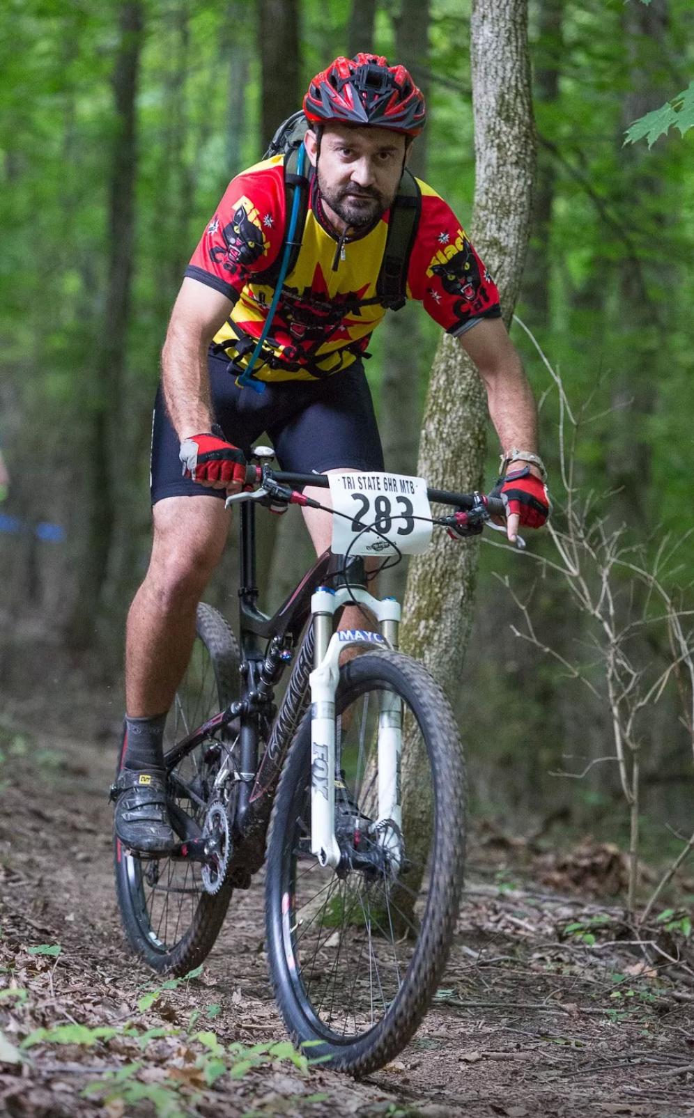 A male mountain biker wearing a brightly colored shirt and a helmet rides along a forest trail. He is focused on navigating the path, surrounded by green trees and foliage. The bike is equipped for off-road terrain, and a race number is visible on the front of his bike. England Idlewild Mountain Biking Park mountain bike trail.