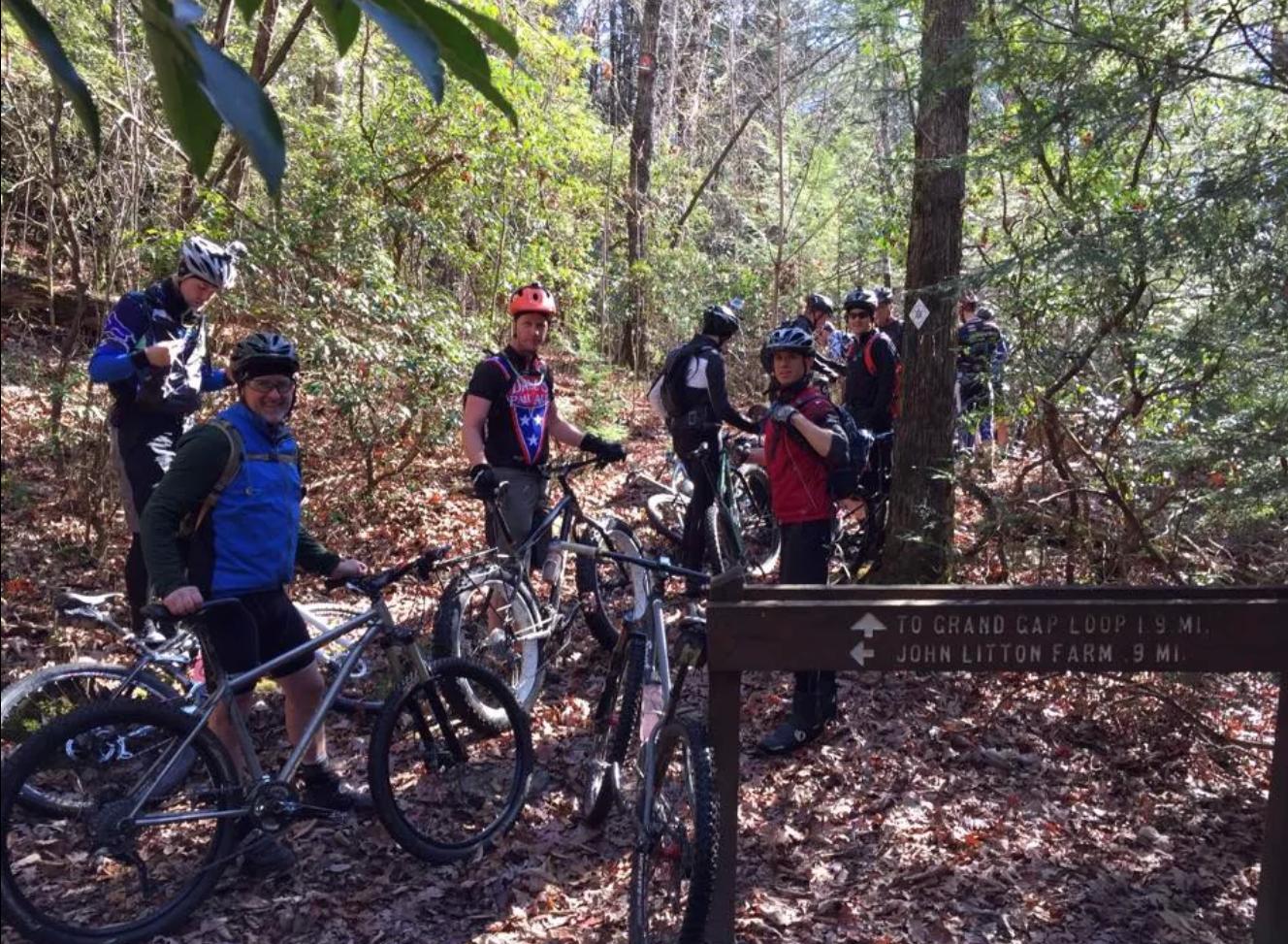 A group of mountain bikers standing near their bikes on a forest trail, surrounded by trees and fallen leaves. A directional sign in the foreground indicates the distances to "Grand Cap Loop" and "John Litton Farm." The bikers are wearing helmets and cycling gear, enjoying a break in the natural setting. Big South Fork mountain bike trail.