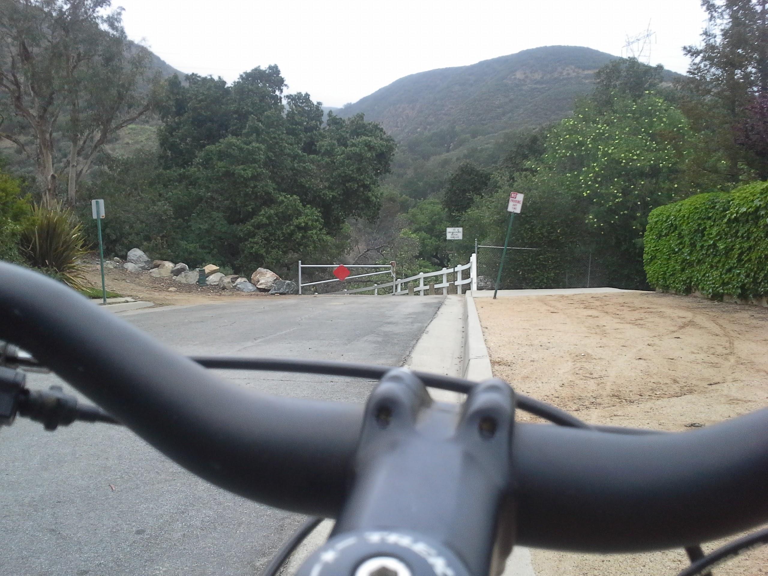 A view from the handlebars of a bicycle, looking down a quiet, winding road leading to a gated area surrounded by greenery and hills. A red stop sign is visible on the gate, and a "No Trespassing" sign can be seen in the background. The setting is peaceful, with trees and a hillside visible, evoking a sense of nature and outdoor adventure. Cucamonga Road mountain bike trail.