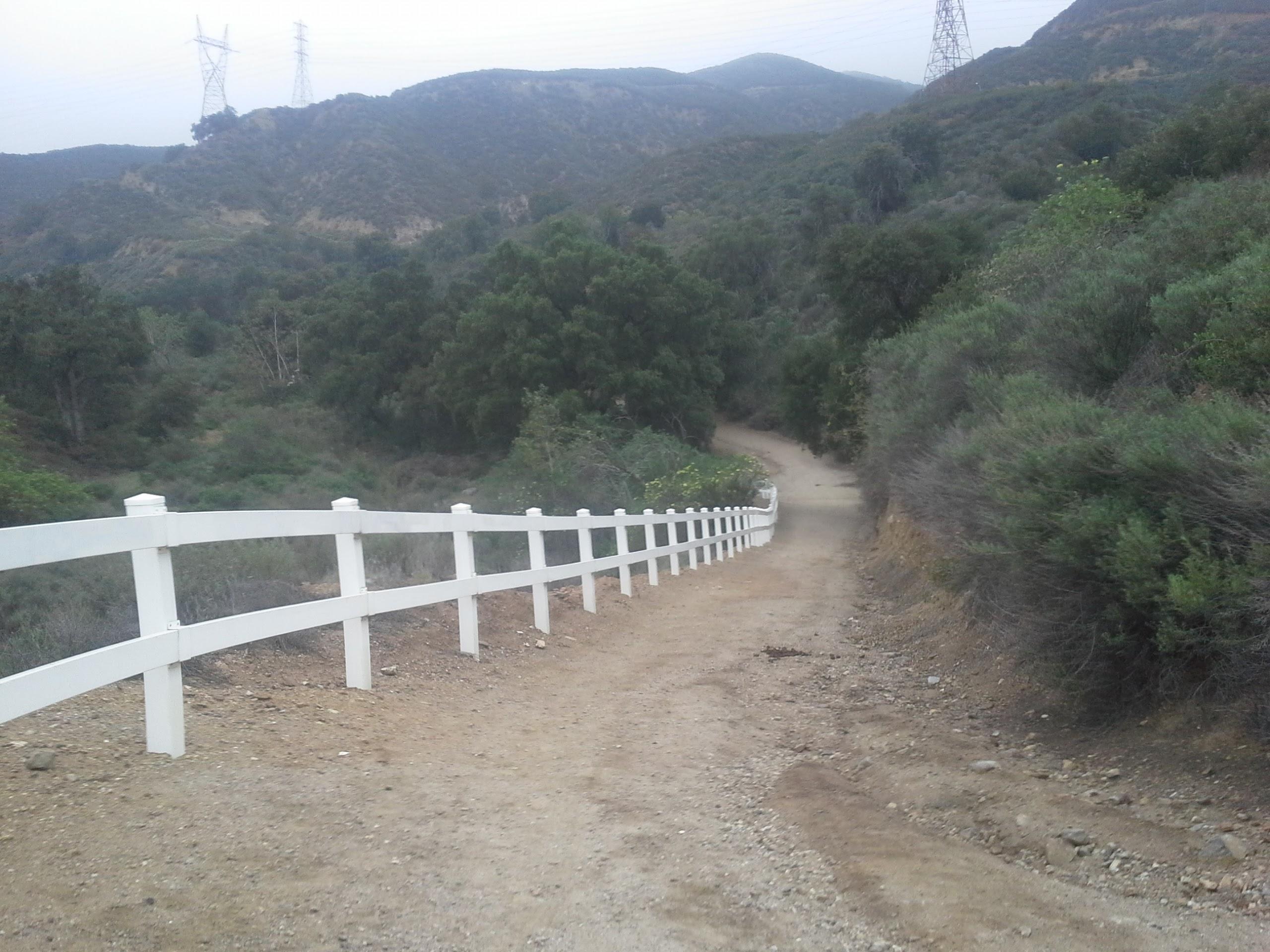 A dirt path winding downhill through a mountainous area, lined with a white fence on one side. Lush greenery and shrubs are present along the path, with power lines visible in the background. The sky is overcast, creating a serene atmosphere in the natural landscape. Cucamonga Road mountain bike trail.