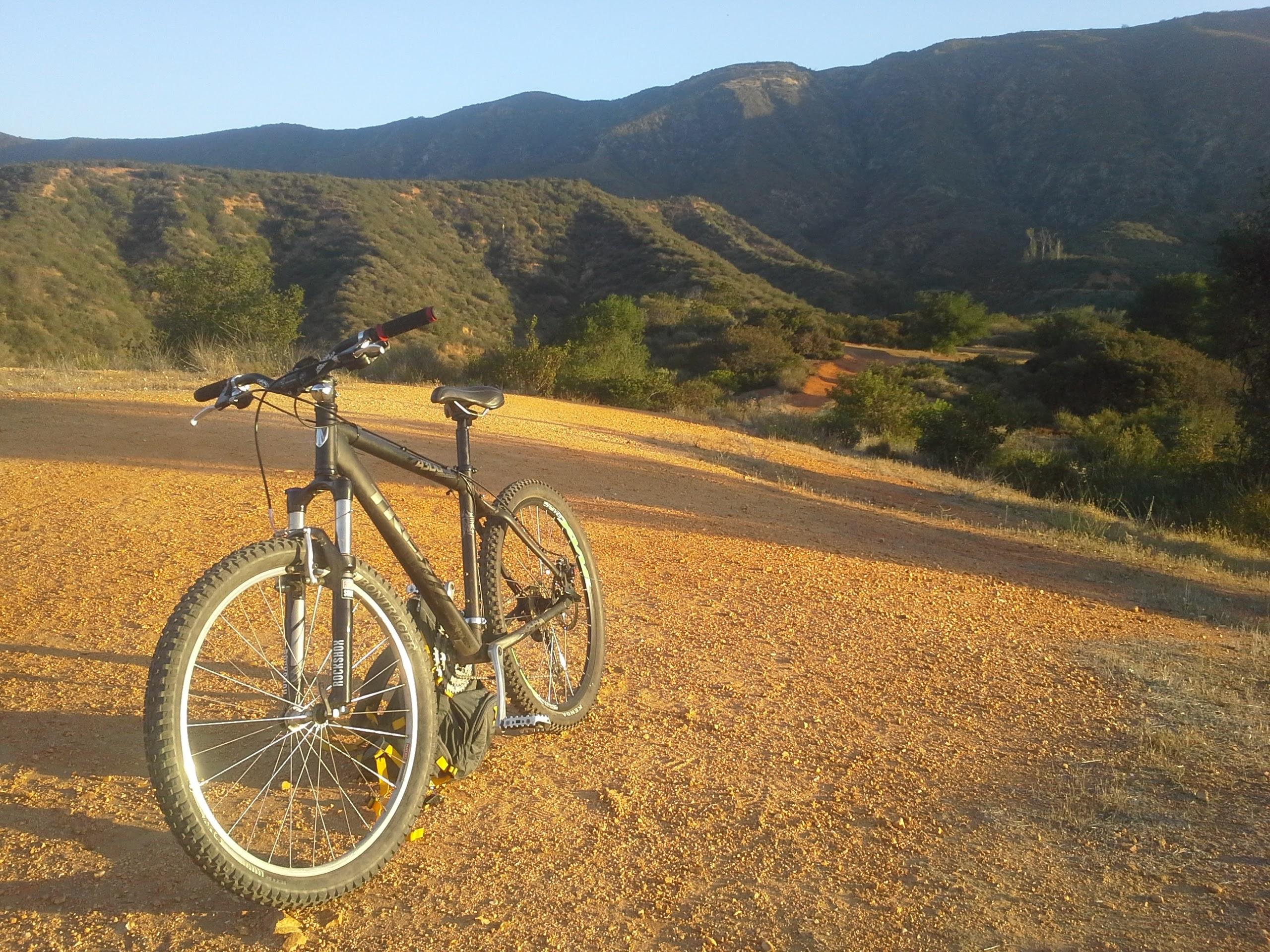 A mountain bike parked on a dirt trail, surrounded by rolling hills and green shrubbery under a clear blue sky. The path is illuminated by sunlight, creating a warm atmosphere in a natural outdoor setting. Claremont Wilderness mountain bike trail.