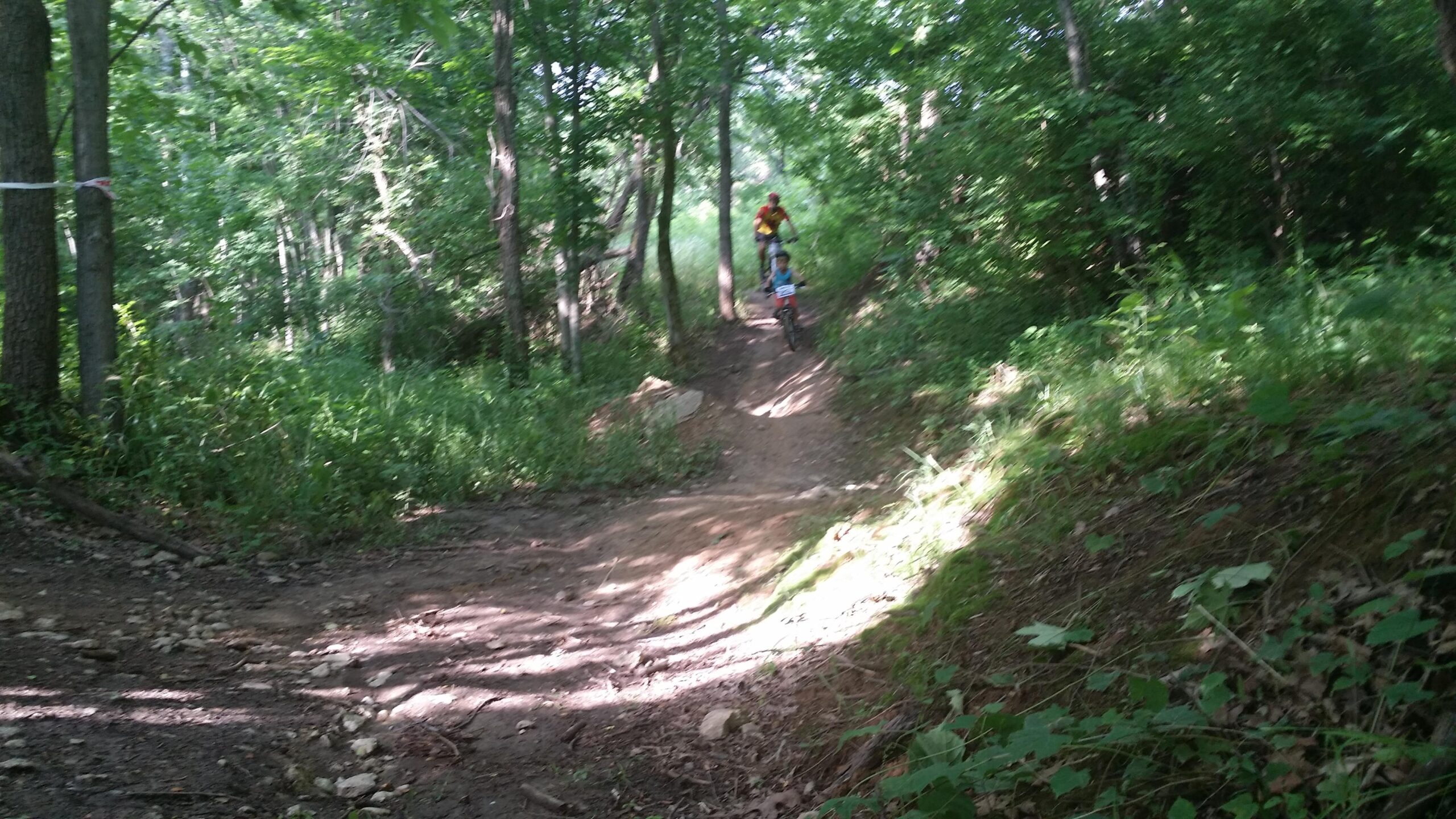 A mountain biker rides along a dirt trail surrounded by greenery in a wooded area, with sunlight filtering through the leaves. The rider is accompanied by another cyclist in the background, both navigating the winding path. England Idlewild Mountain Biking Park mountain bike trail.