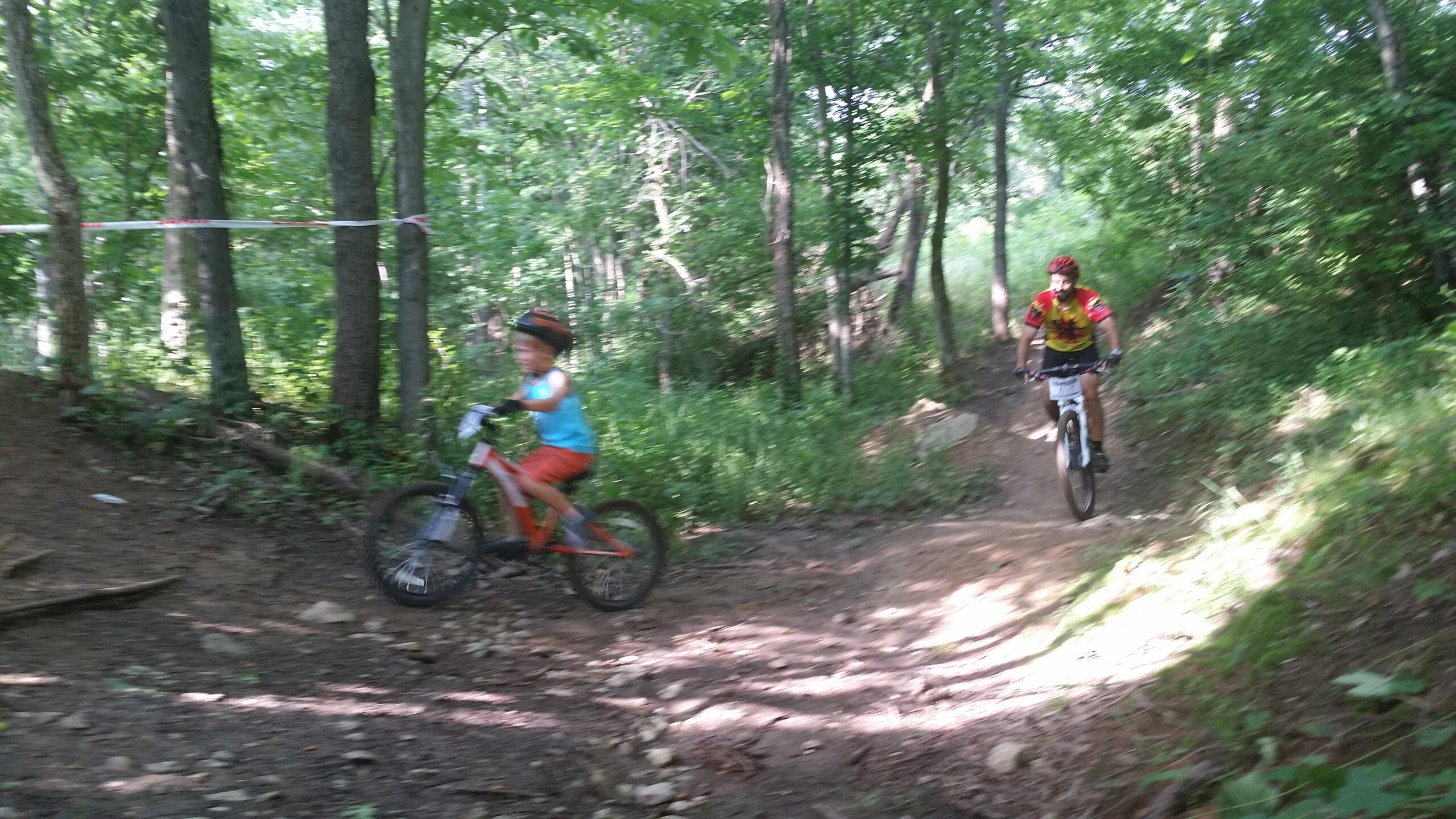 A child riding a bicycle along a dirt trail in a wooded area, while an adult cyclist approaches from behind. The scene is sunny with green trees and foliage surrounding the path, creating a lively outdoor atmosphere. England Idlewild Mountain Biking Park mountain bike trail.