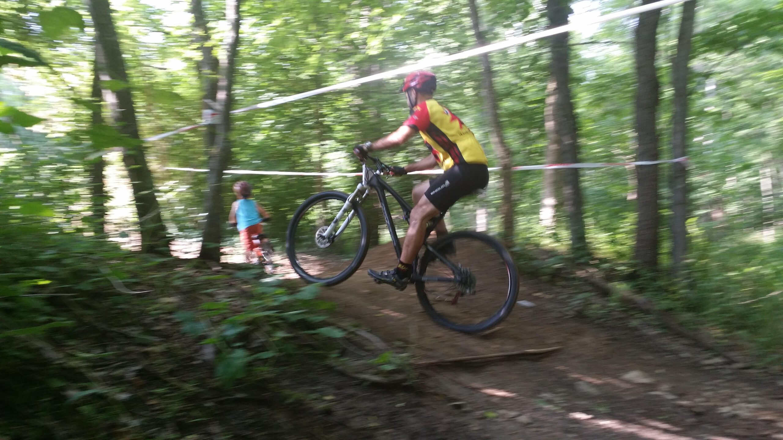 A mountain biker performing a wheelie on a wooded trail, with green trees surrounding the path. In the background, a child is watching the biker, dressed in casual clothing. The image has a slight motion blur, conveying the action of the biker in mid-movement. England Idlewild Mountain Biking Park mountain bike trail.