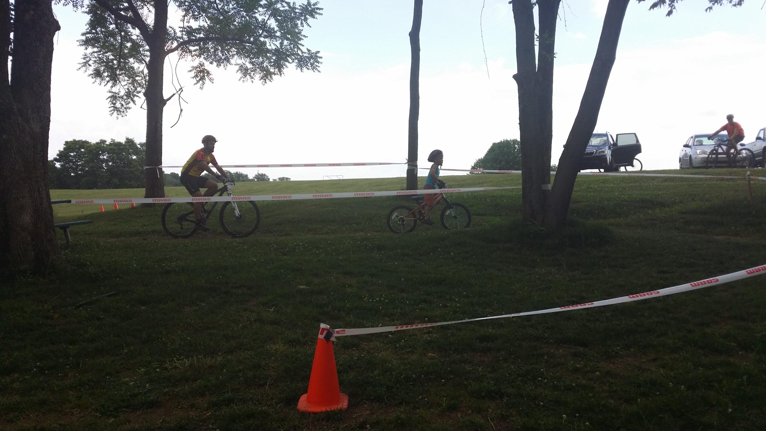 A man and a young child riding bicycles on a grassy area surrounded by trees, with a course marked by safety tape. There are traffic cones positioned nearby, and several cars are parked in the background. England Idlewild Mountain Biking Park mountain bike trail.