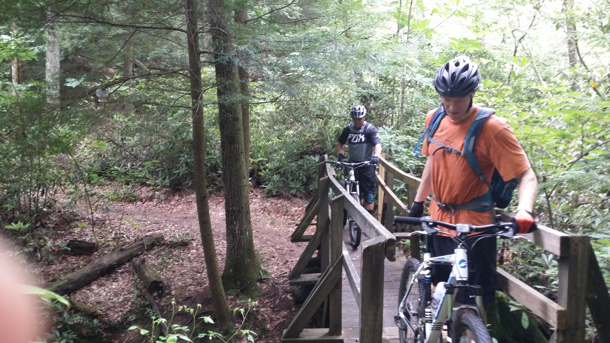 Two mountain bikers navigating a wooden bridge in a lush, green forest. One rider, wearing an orange shirt and a backpack, is looking down at his bike, while the other, dressed in a black shirt and helmet, stands nearby. The surroundings are filled with trees and foliage, creating a natural, outdoor atmosphere. Big South Fork mountain bike trail.