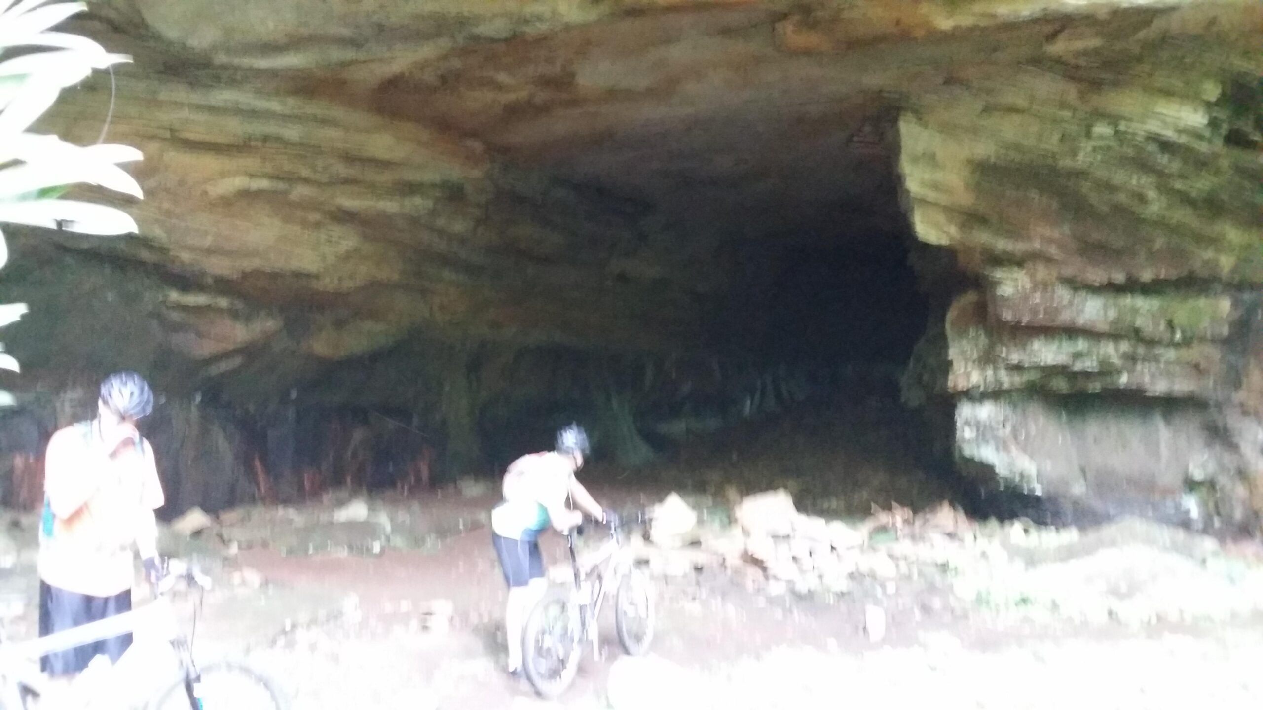 Two mountain bikers near a cave entrance, surrounded by natural rock formations and vegetation. One biker is adjusting their bike while the other appears to be taking a break, with the dark interior of the cave visible in the background. Big South Fork mountain bike trail.