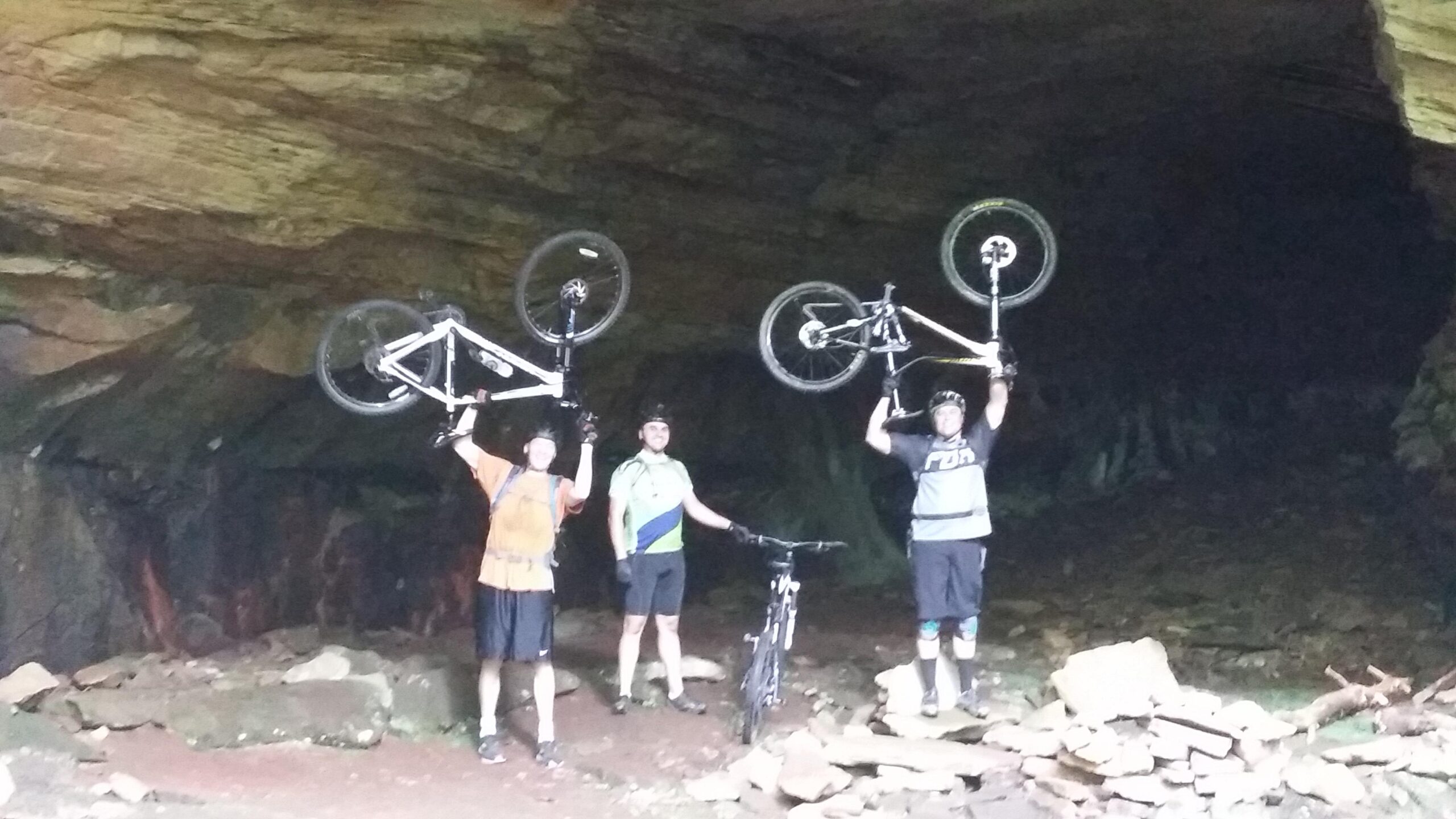 Three mountain bikers stand inside a cave, each holding their bikes above their heads. The background features rough, rocky walls, and scattered stones on the ground. The bikers are wearing helmets and biking gear, smiling proudly at the camera. Big South Fork mountain bike trail.