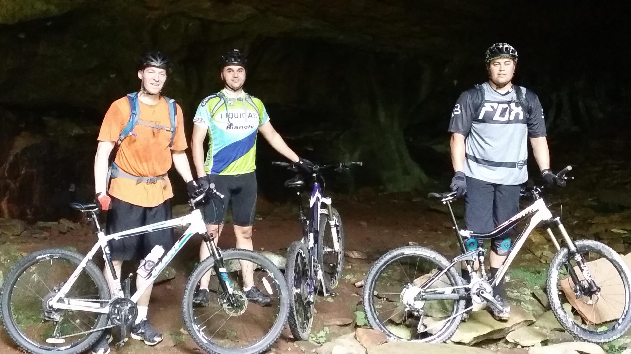 Three mountain bikers posing with their bikes inside a rocky cave. The individuals are wearing helmets and cycling gear, and the environment is dimly lit, featuring rough terrain and rock formations in the background. Big South Fork mountain bike trail.