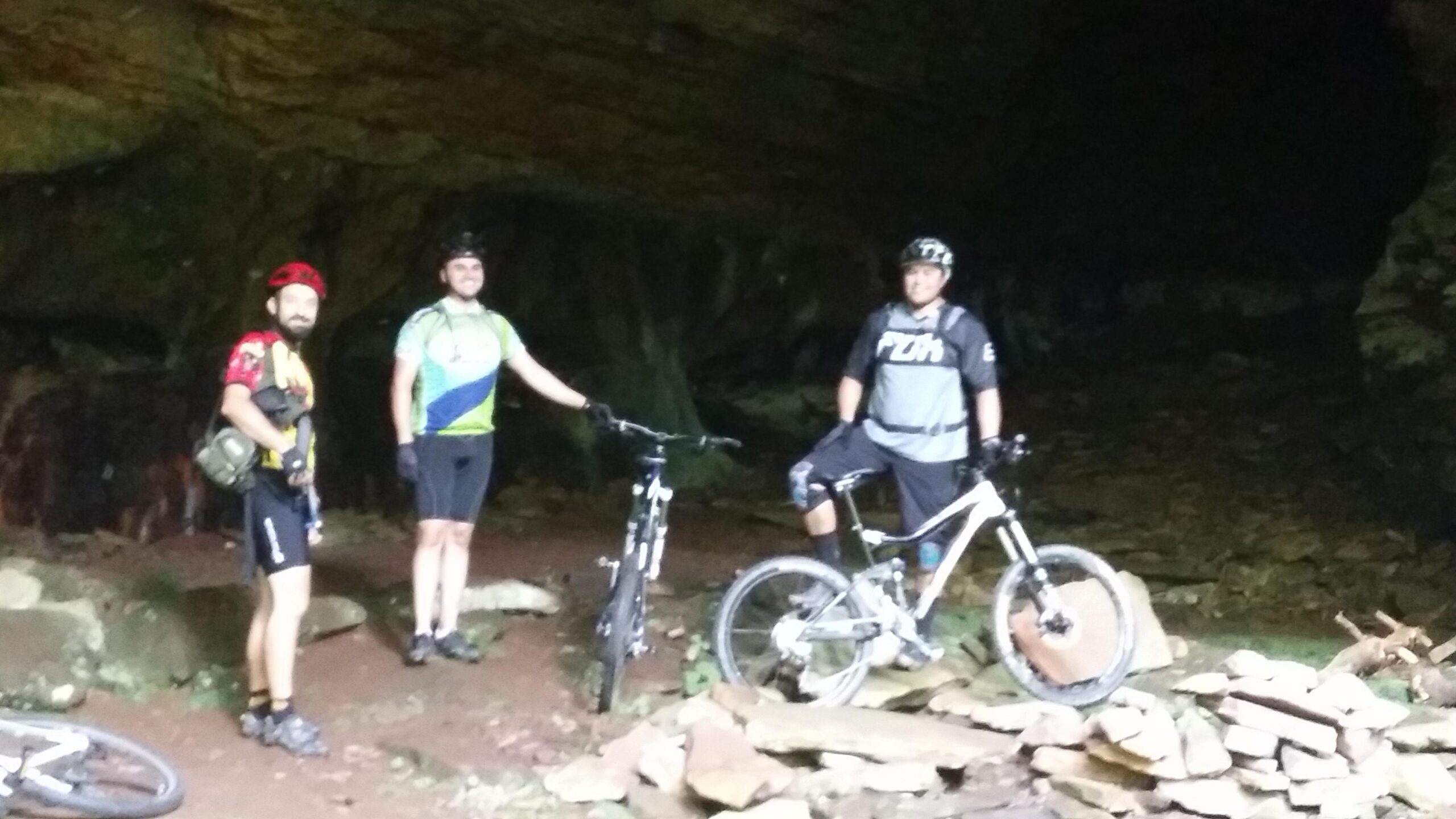 Three mountain bikers standing inside a cave, surrounded by rock formations. They are wearing cycling gear and helmets, with their bicycles nearby. The atmosphere is dimly lit, showcasing the cave's natural features. Big South Fork mountain bike trail.