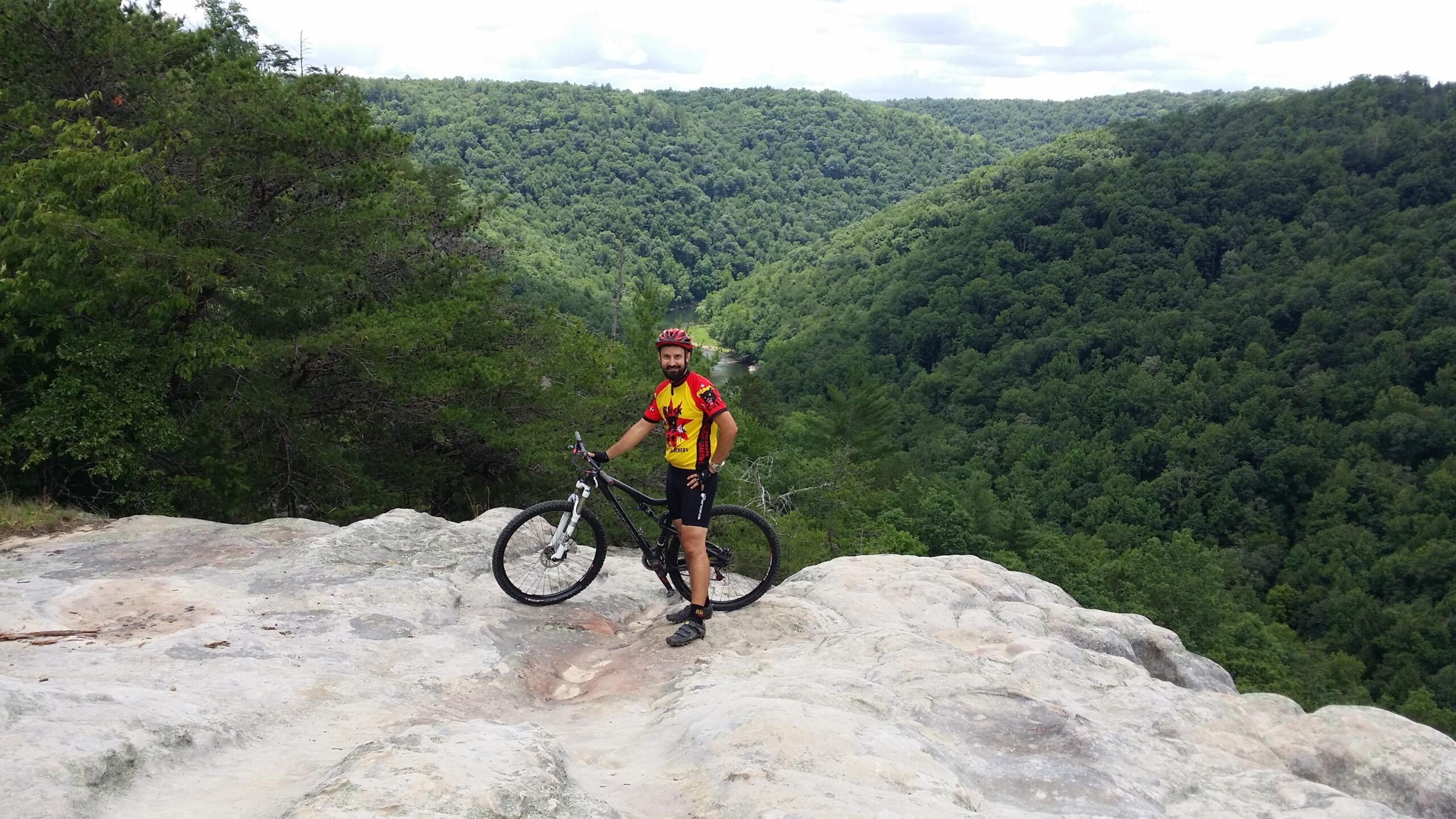 A male mountain biker stands on a rocky outcrop, with a lush green valley and hills in the background. He wears a colorful cycling jersey and helmet, and is next to his mountain bike. The scene is set on a clear day with scattered clouds, showcasing a panoramic view of the surrounding forested landscape. Big South Fork mountain bike trail.