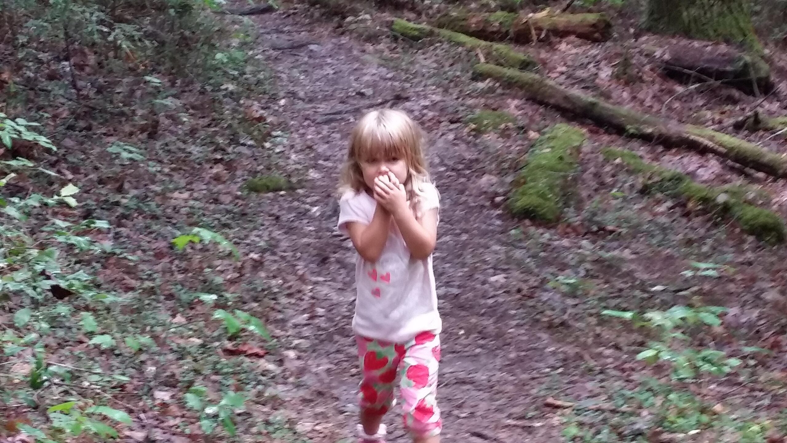 A young child with blonde hair is walking along a narrow, muddy trail in a forest. She is wearing a light-colored t-shirt with small heart patterns and pink pants decorated with larger heart shapes. The child appears to be playfully covering her mouth with her hands, surrounded by greenery and fallen leaves on the ground. Cane Creek (sheltowee Trace Trail) mountain bike trail.