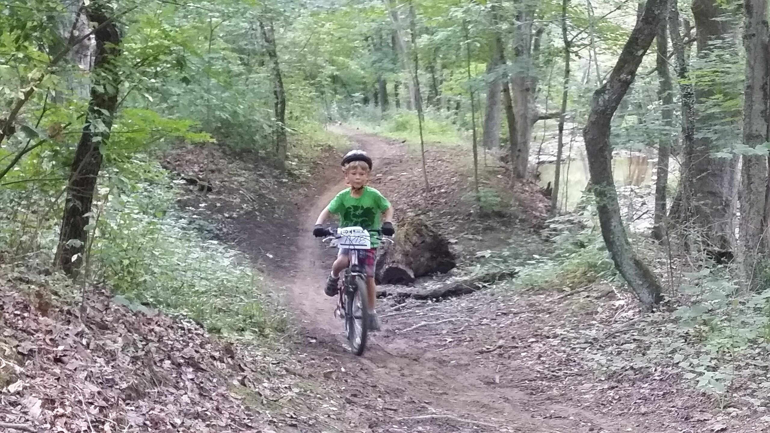 A young boy riding a mountain bike on a dirt trail through a wooded area, surrounded by lush green foliage and trees. England Idlewild Mountain Biking Park mountain bike trail.