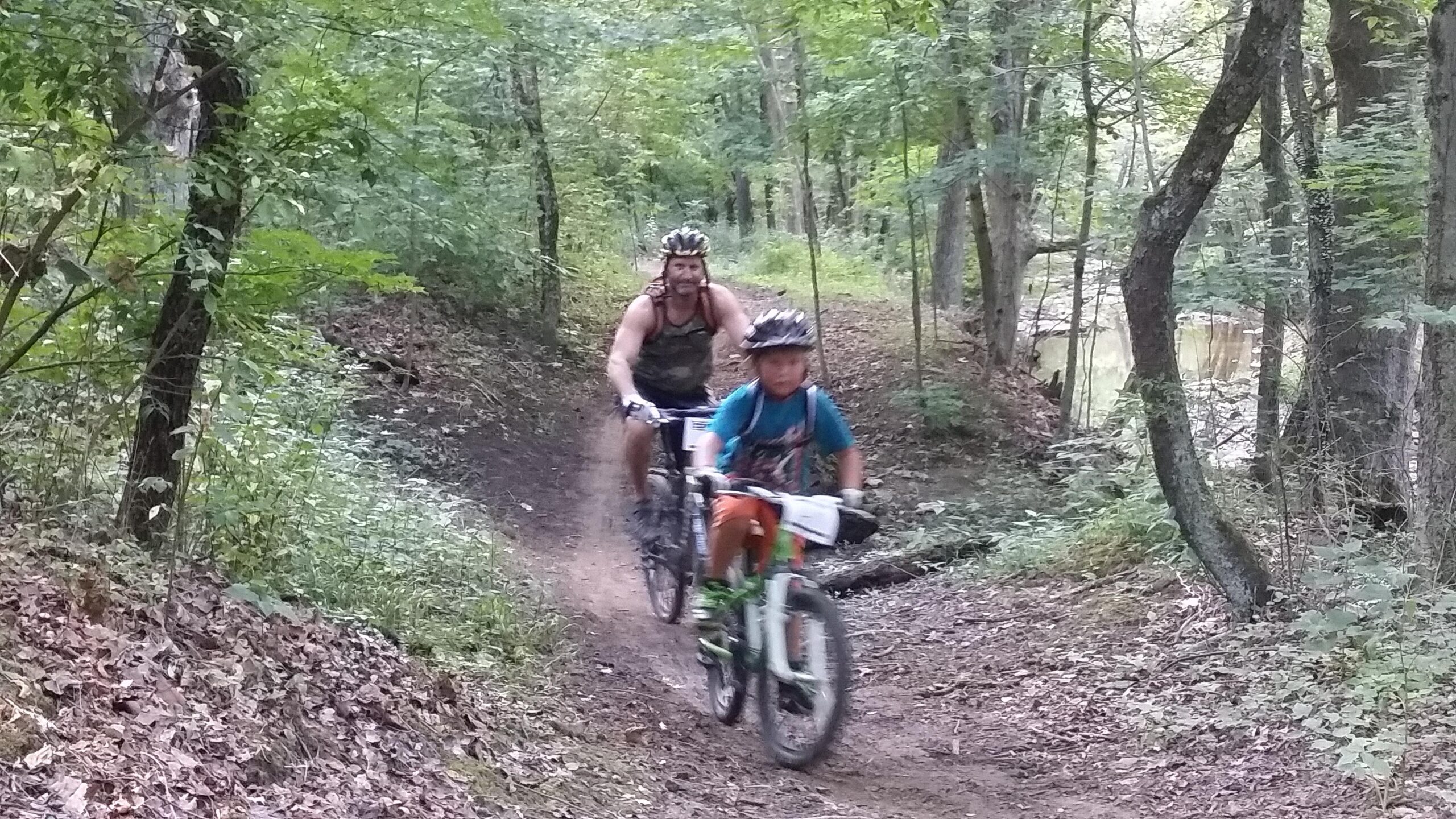 A man and a young boy ride mountain bikes along a dirt trail in a wooded area, surrounded by green trees and foliage. The boy is in the foreground, wearing a blue shirt and orange shorts, while the man follows closely behind. The scene captures a moment of outdoor adventure and enjoyment in nature. England Idlewild Mountain Biking Park mountain bike trail.