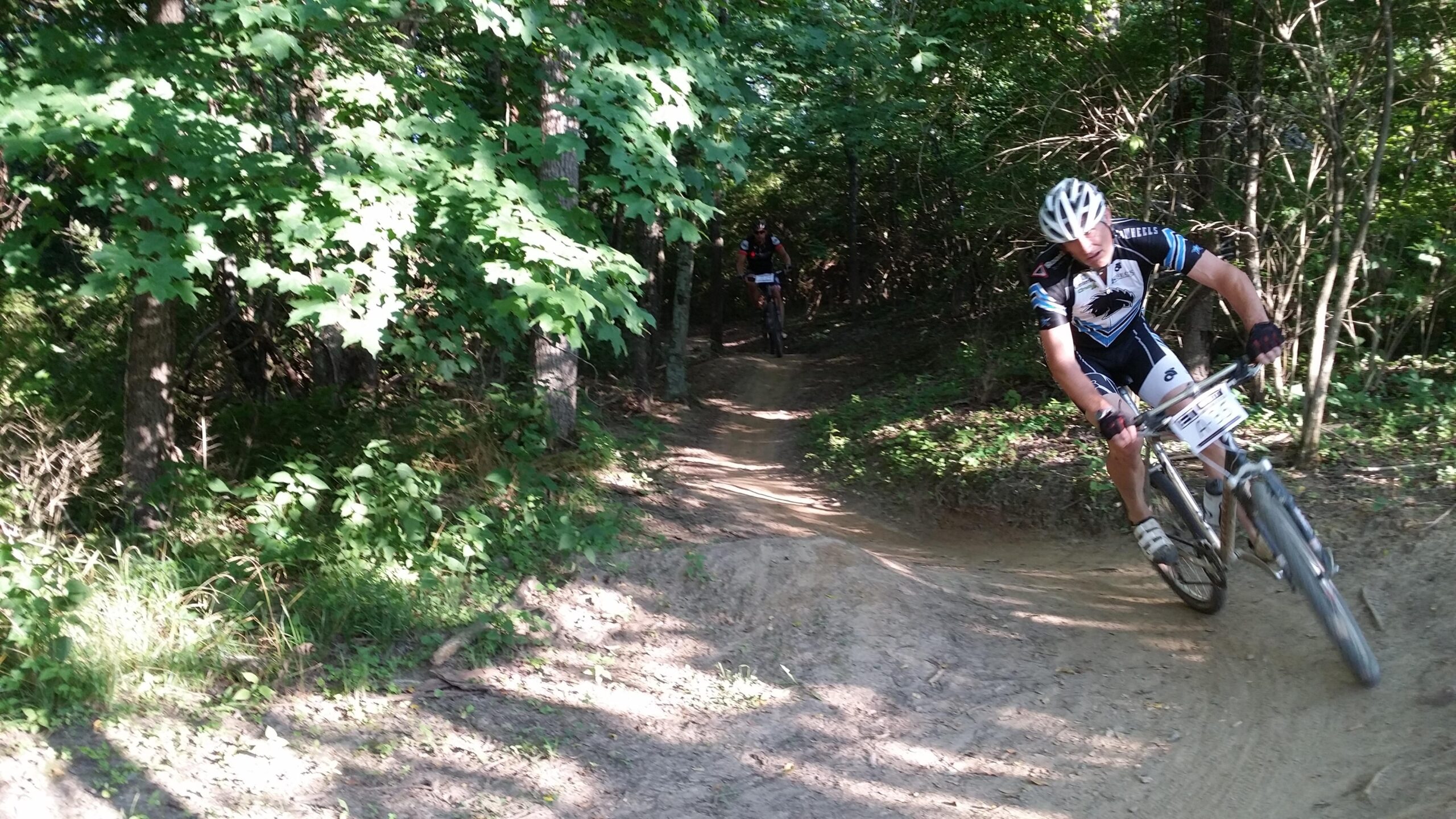 A mountain biker navigating a dirt trail through a wooded area, surrounded by greenery and trees. The rider appears to be focused, leaning into a turn as they navigate the course. A second cyclist is visible in the background, also riding along the trail. England Idlewild Mountain Biking Park mountain bike trail.