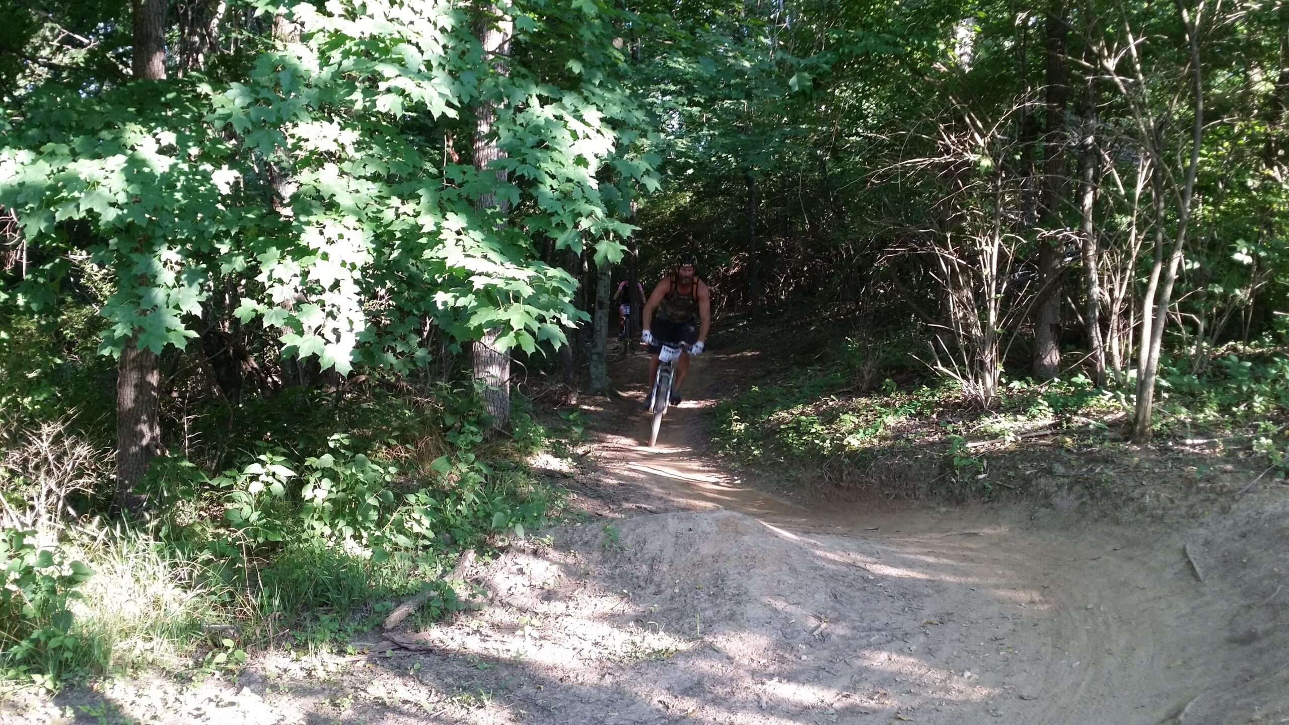 A person riding a mountain bike on a narrow dirt trail surrounded by lush green trees and foliage. England Idlewild Mountain Biking Park mountain bike trail.