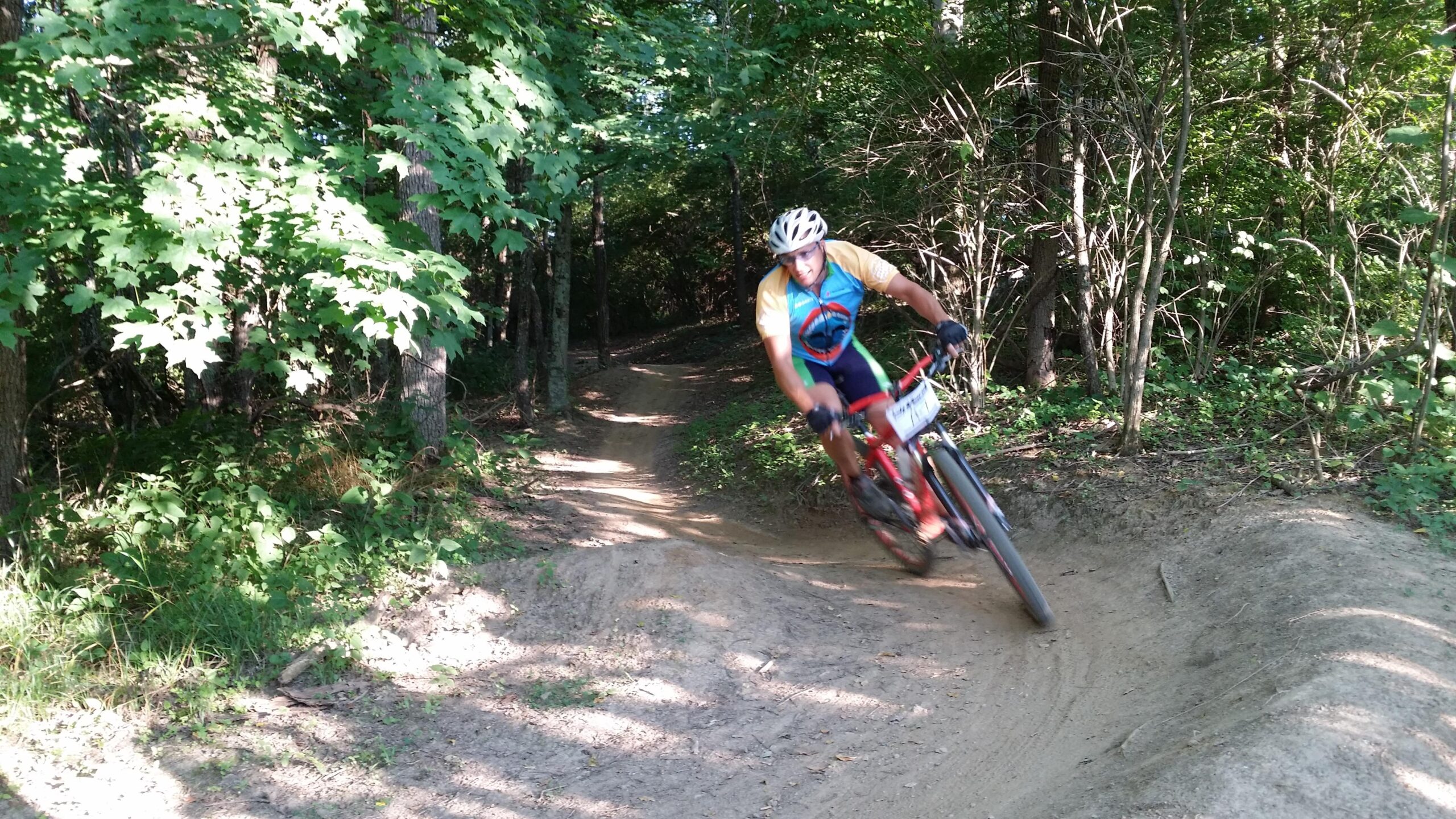 A mountain biker navigating a dirt trail in a forested area, leaning into a turn. The cyclist is wearing a helmet and colorful cycling attire, with green trees and underbrush in the background. Sunlight filters through the leaves, creating a vibrant outdoor scene. England Idlewild Mountain Biking Park mountain bike trail.