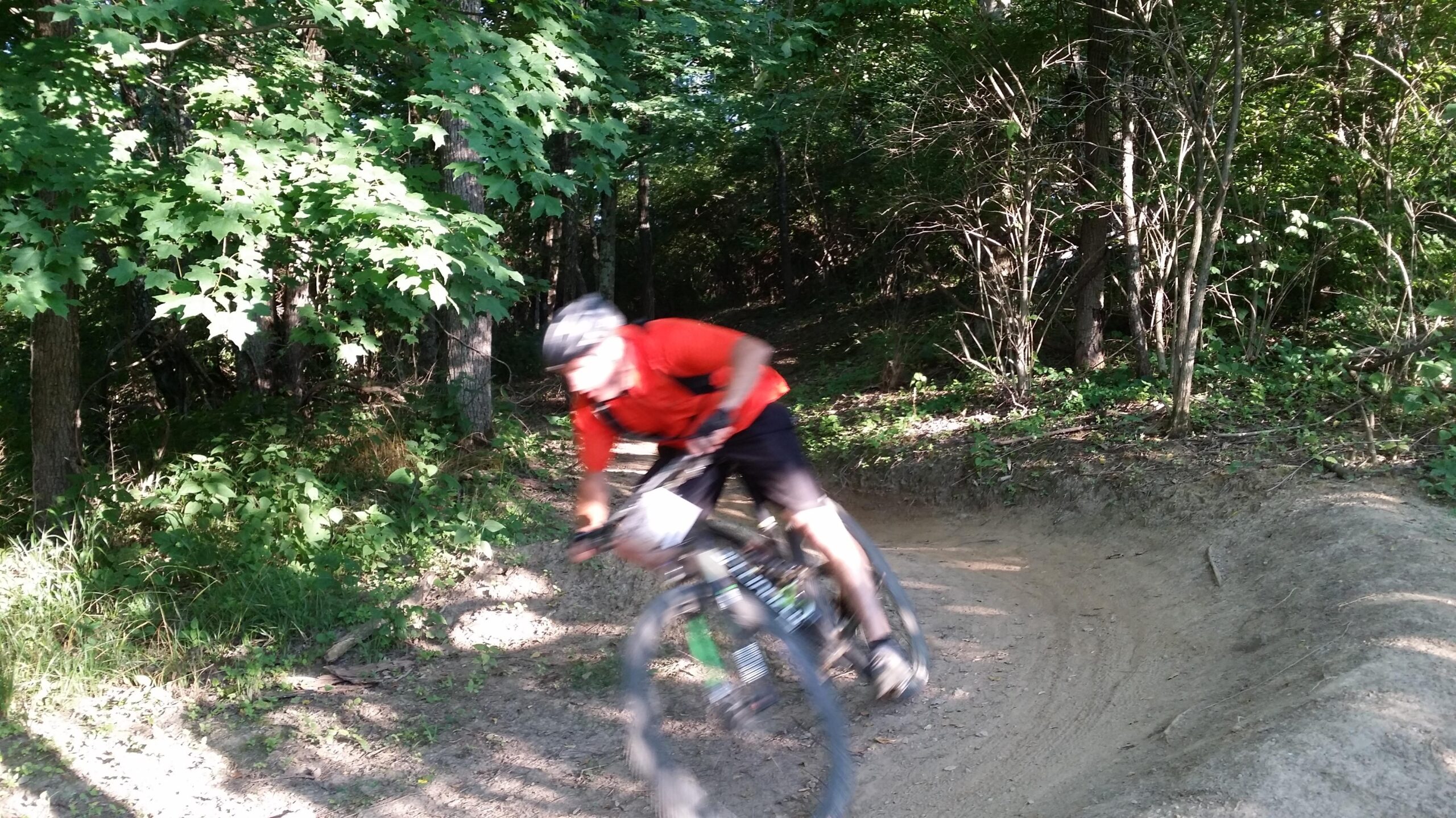 A mountain biker in a red shirt leans into a turn on a dirt trail surrounded by lush green trees and underbrush, with motion blur indicating speed and movement. England Idlewild Mountain Biking Park mountain bike trail.